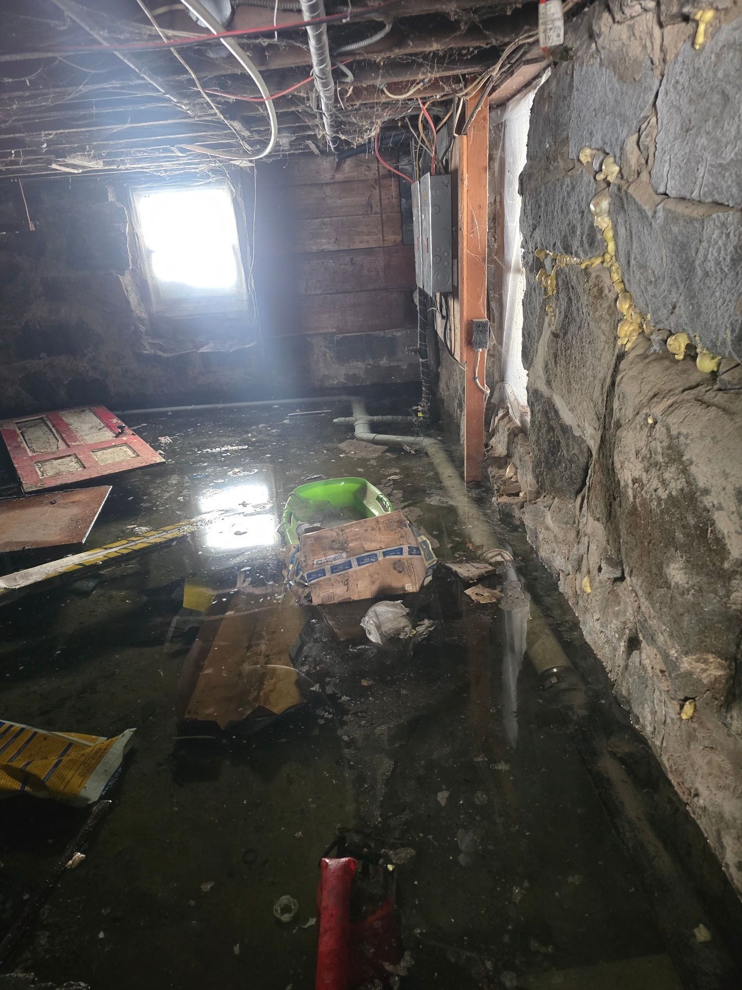 Flooded basement with standing water, debris, exposed stone walls, and a small window.