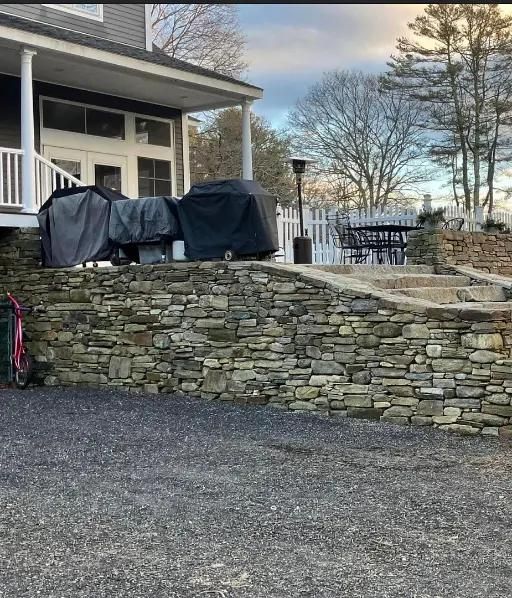 Stone retaining wall in front of a house with covered grills on a patio, stairs, and a white fence.