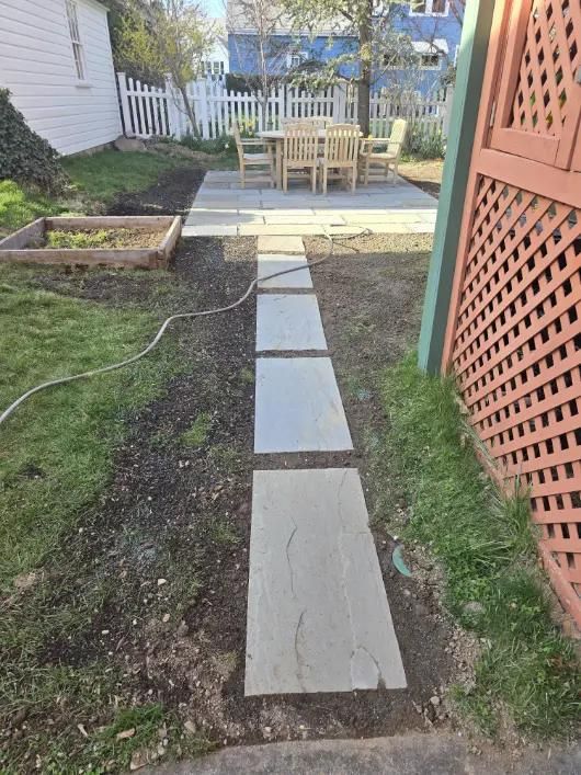 Path of gray stone pavers leading to a patio with a wooden table and chairs, surrounded by grass and a garden.