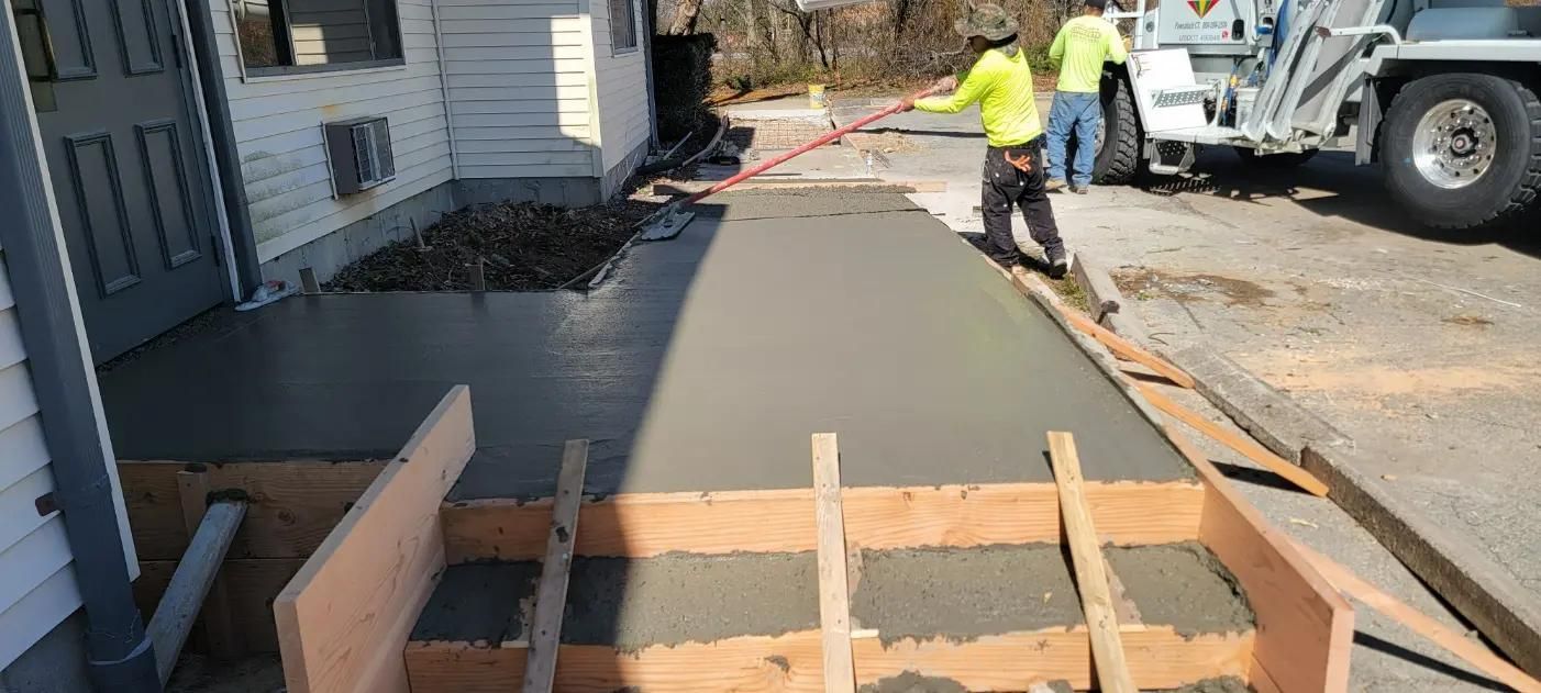 Workers pouring and smoothing fresh concrete for a sidewalk next to a building.