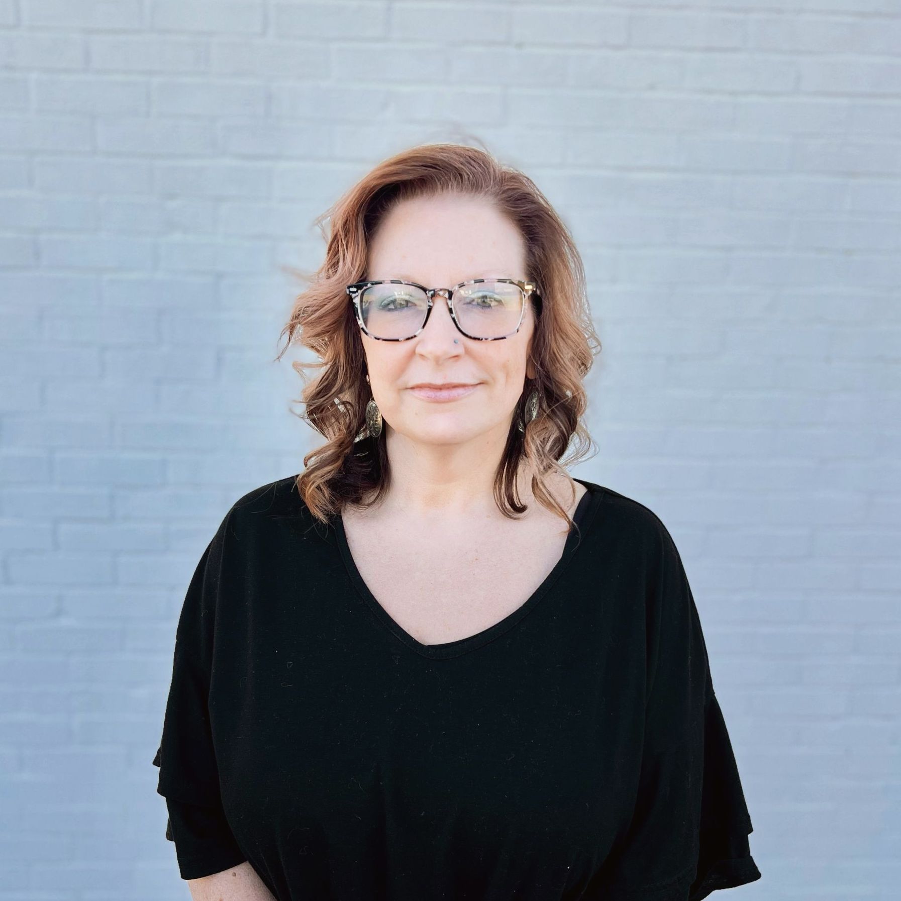 A woman wearing glasses and a black shirt is standing in front of a white brick wall.