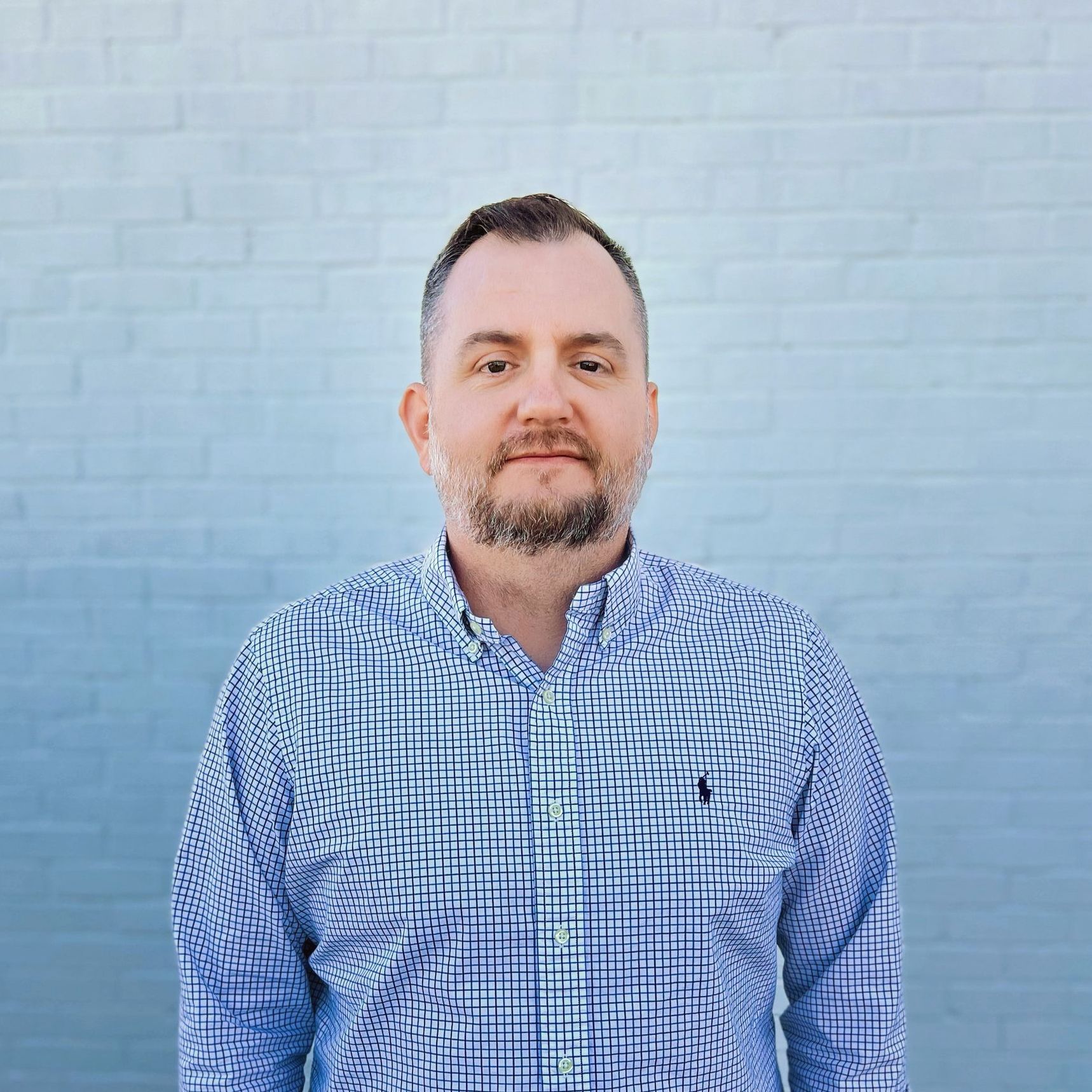 A man with a beard is wearing a blue shirt and standing in front of a blue brick wall.