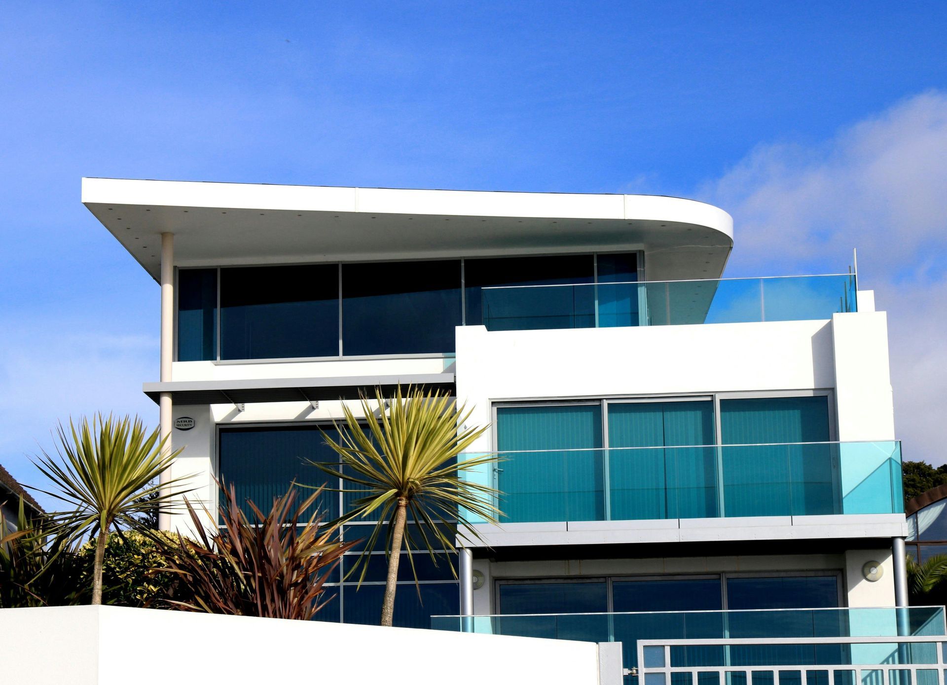 A modern, three-story white house with large glass windows, balconies, and palm trees under a bright blue sky.