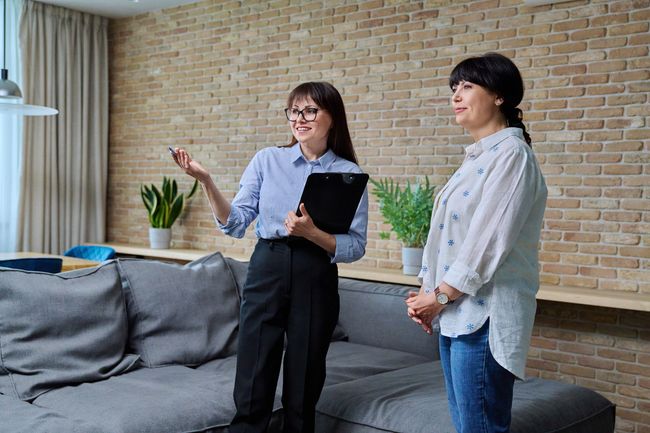 Two people standing in a living room, one holding a tablet and gesturing while the other listens.