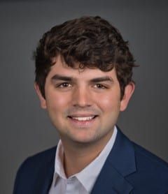 A smiling young man with brown hair wearing a white shirt and navy blue blazer against a gray background.