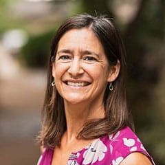 A woman with shoulder-length brown hair smiles outdoors. She wears a floral pink top and small silver earrings.