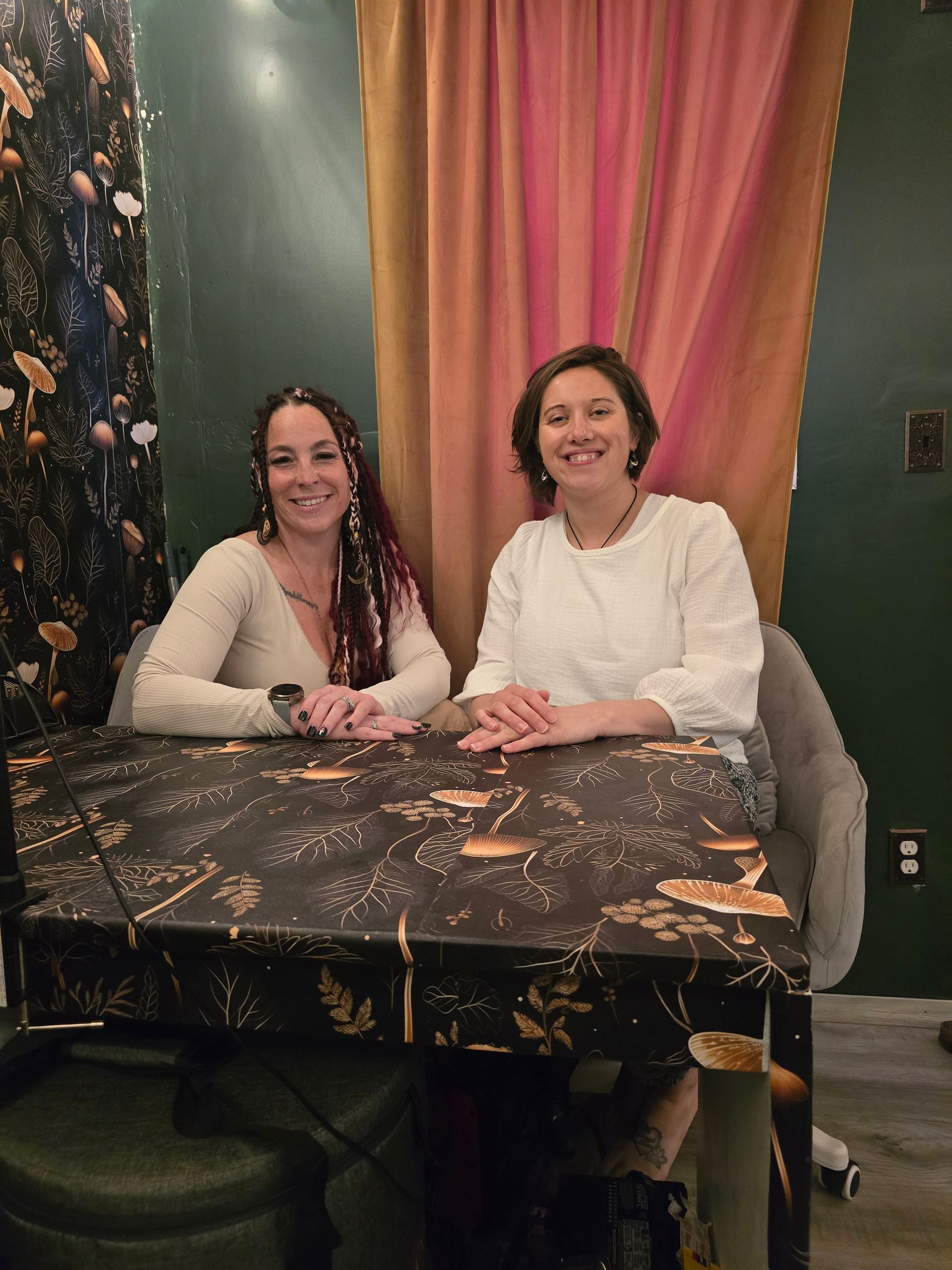 Two women sitting at a desk in front of a gold window curtain.