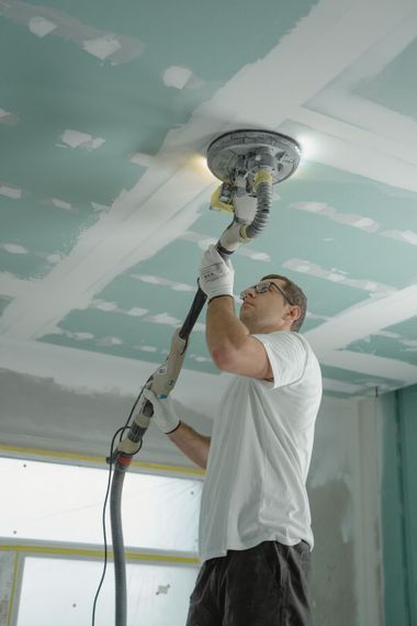 Man using a drywall sander on a ceiling with green primer patches in a room under renovation
