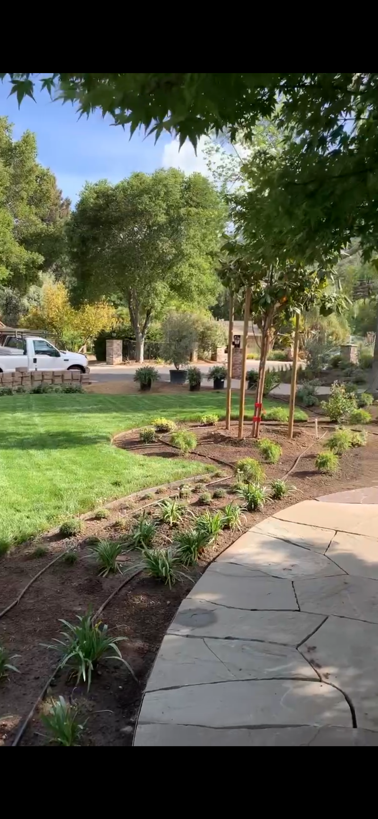 Park landscape with trees, green grass, a paved path, and planted areas. A white truck is visible.