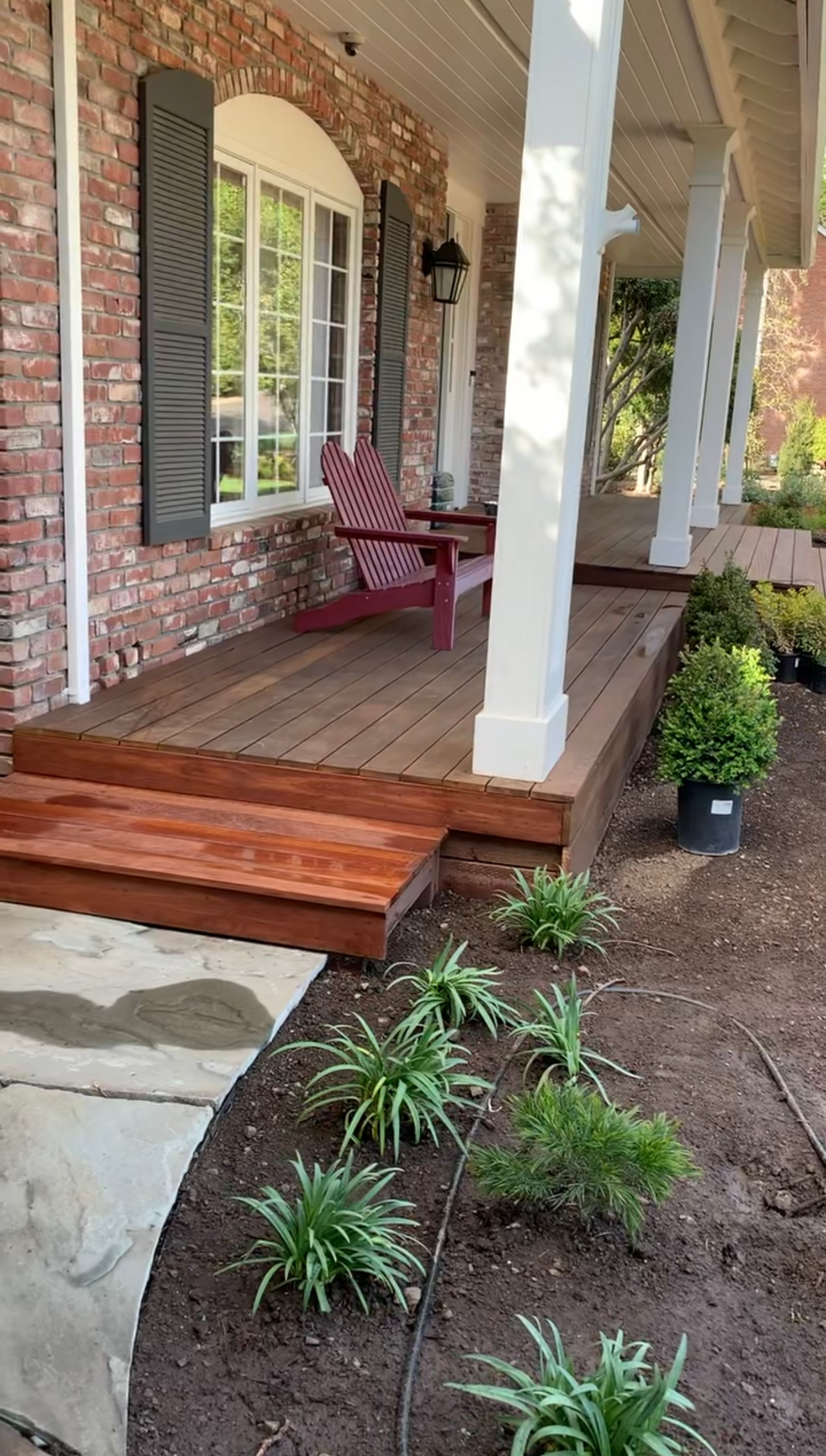 Red-brick house with a wooden porch, steps, and a row of plants. A red chair sits on the porch.