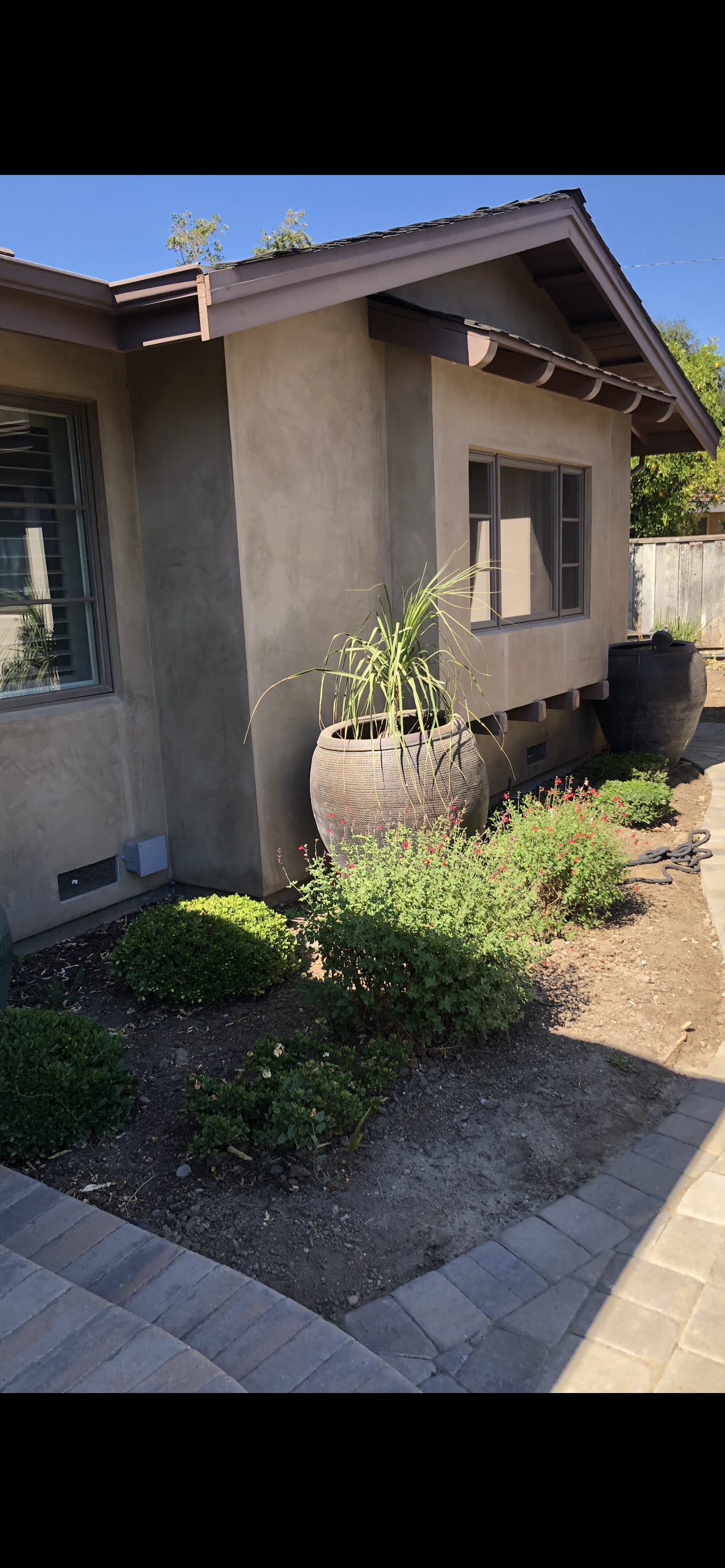 A tan stucco house with a flower bed in front. A large wooden pot has plants. Sunny day.