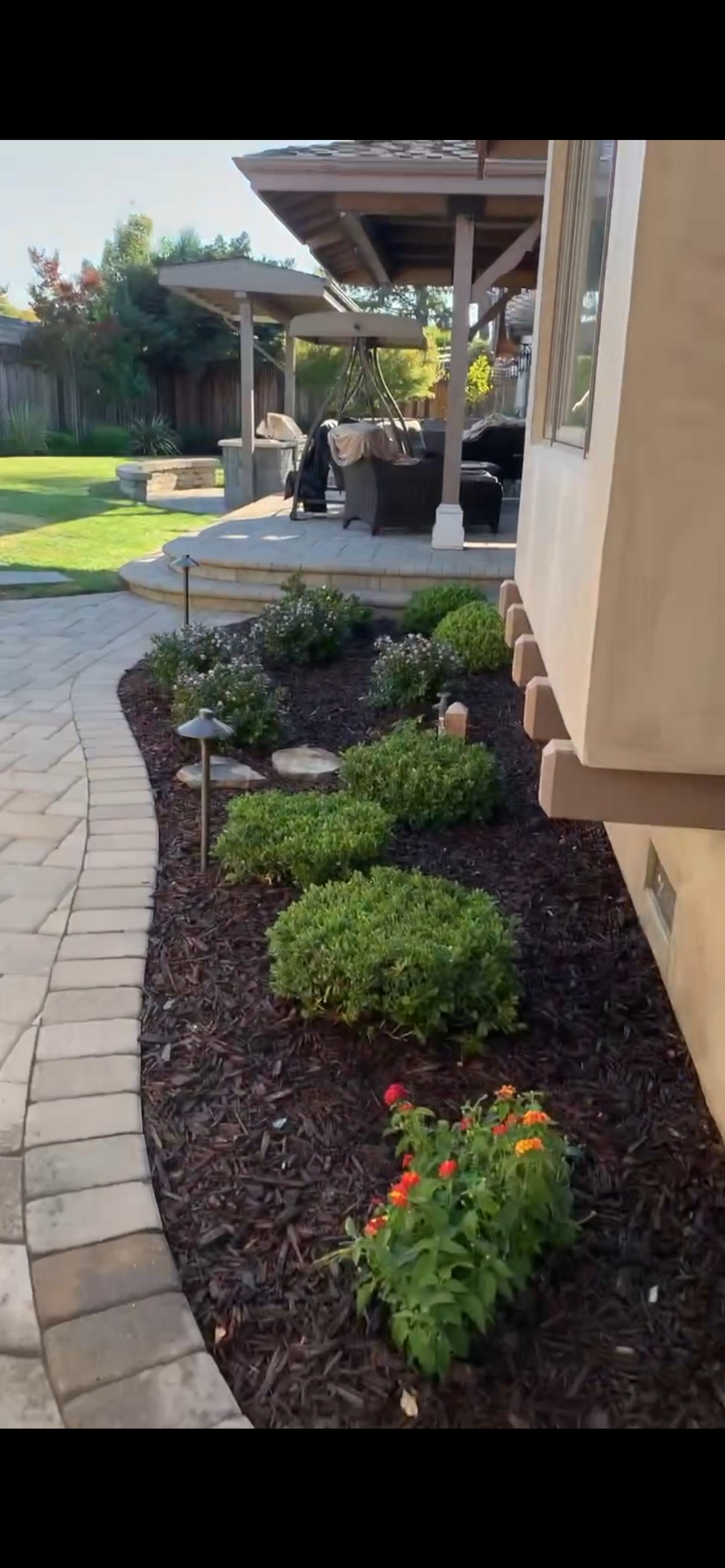 Garden bed with lush green plants, brick pathway, and a patio with a pergola.