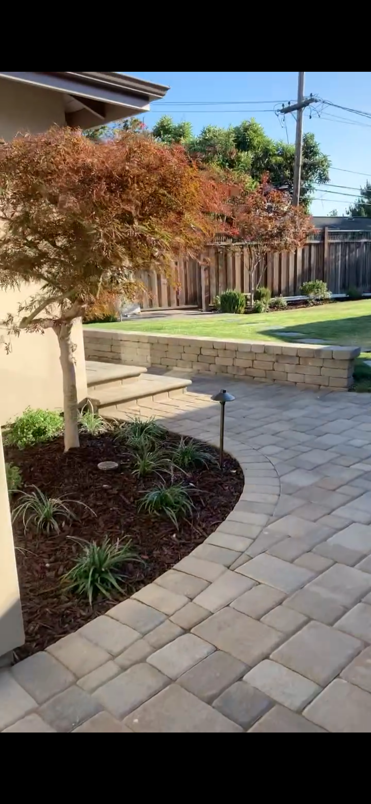 A landscaped backyard with a stone path, retaining wall, and tree with red leaves.