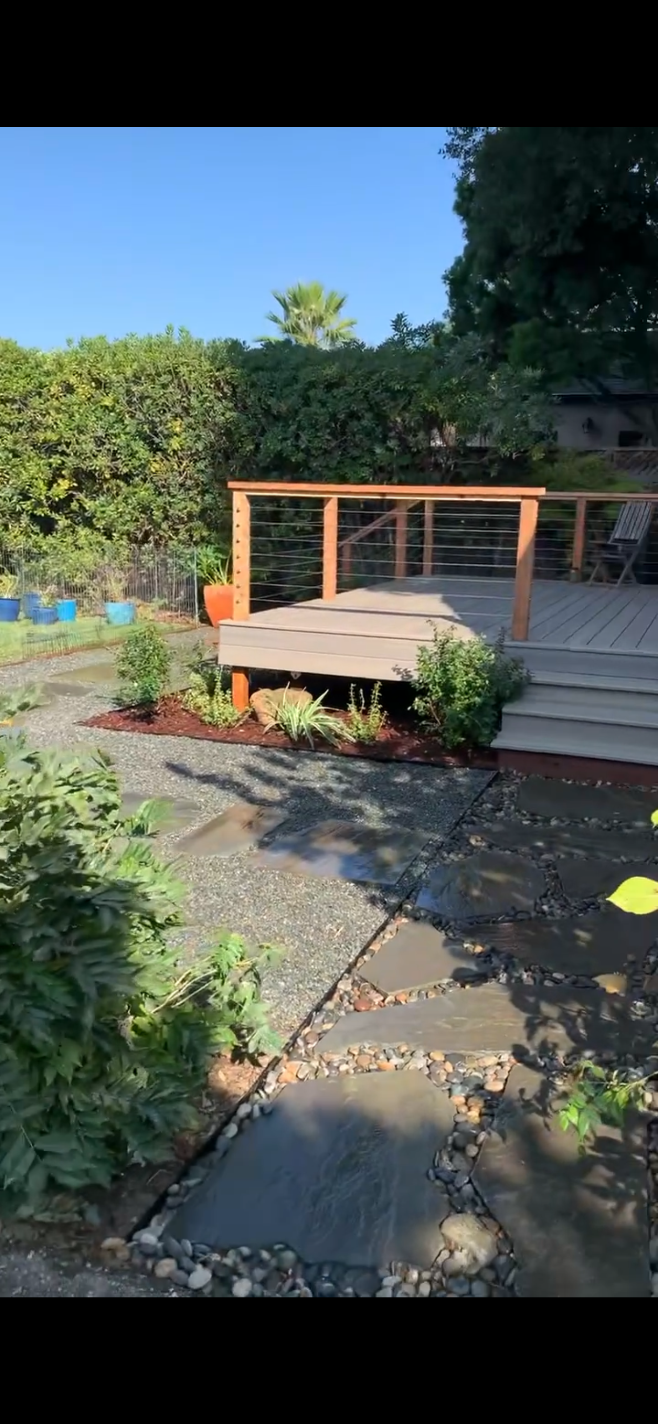 A backyard with a wooden deck, a gravel path, and lush greenery under a clear blue sky.