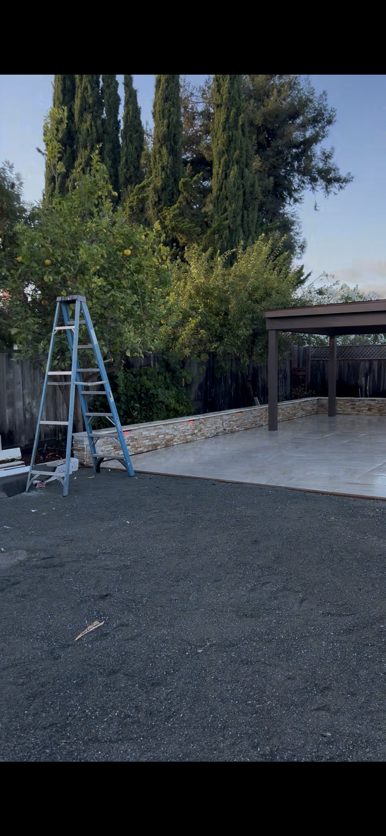 A-frame ladder next to a stone wall and gravel ground, with a patio and trees in the background.