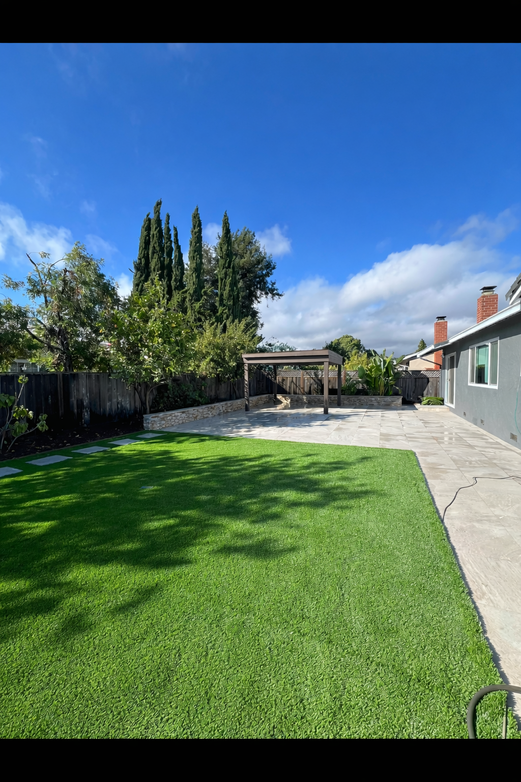 Backyard with green lawn, stone patio, wooden pergola, trees, and blue sky.