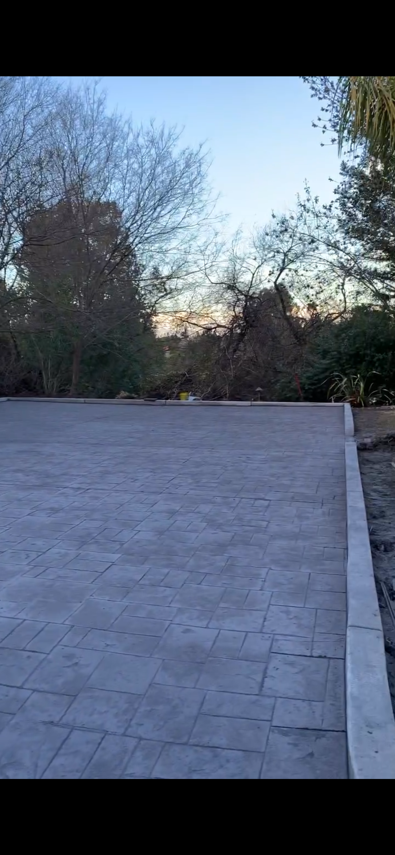Grey paved area with a border, and trees in the background under a blue and orange sky.
