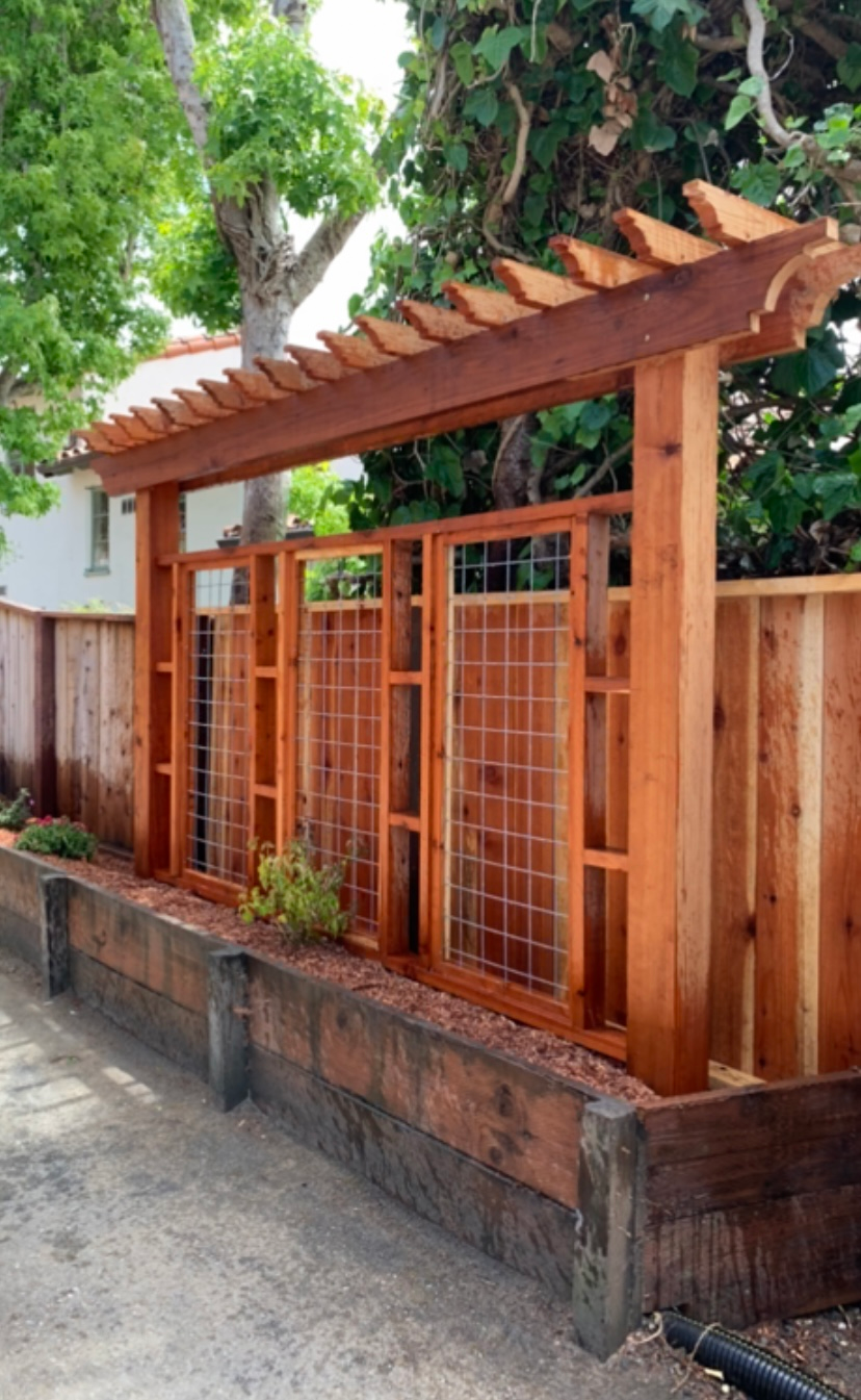 Wooden arbor with wire mesh panels, set in a wood and stone planter, next to a wooden fence.