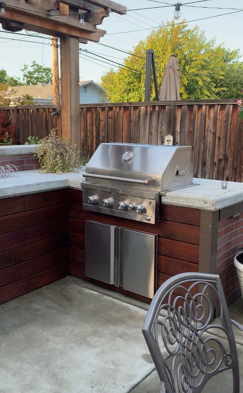 Outdoor built-in grill with stainless steel features, wooden accents, and a brick base in a backyard setting.