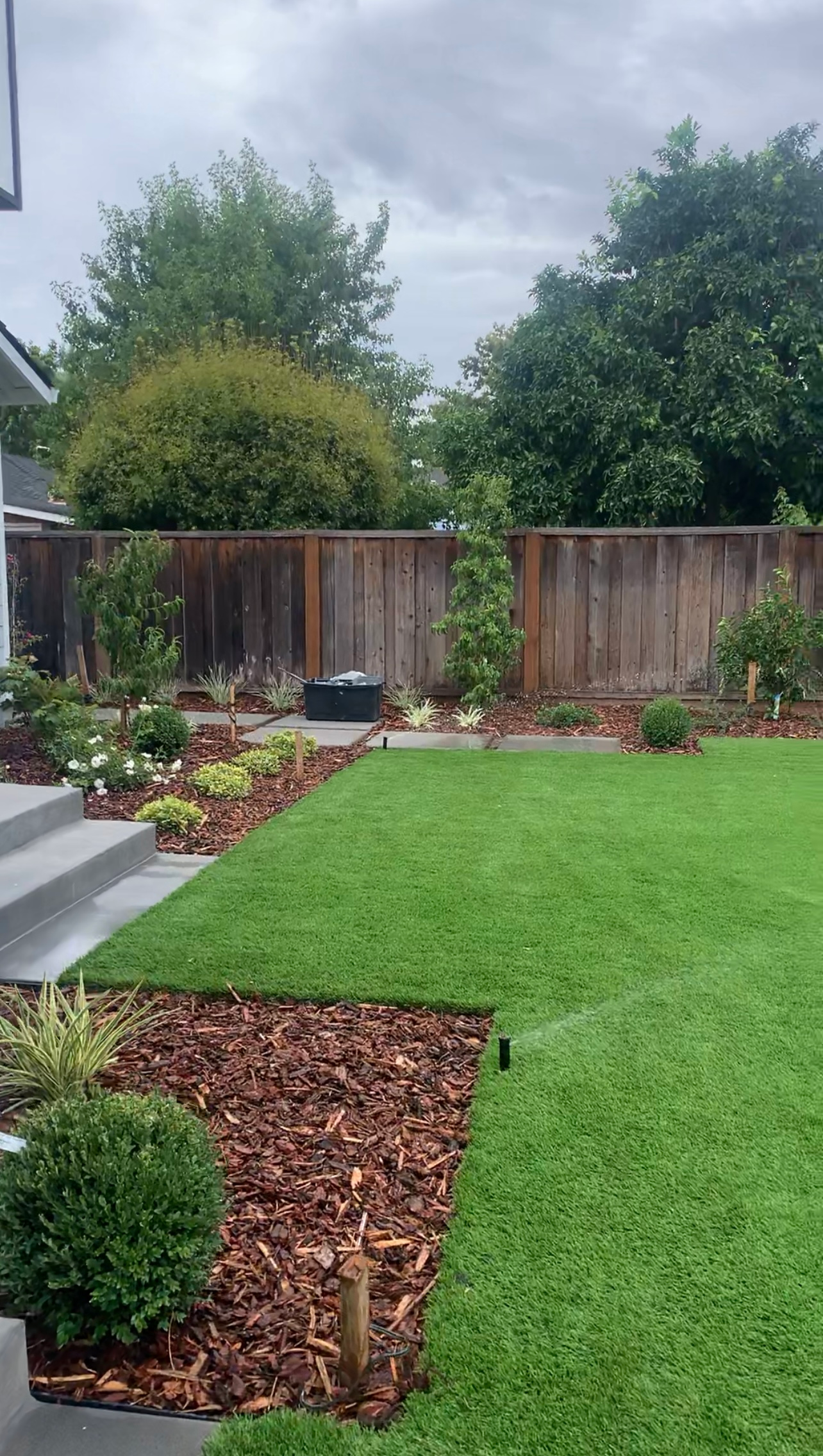 A well-manicured lawn and garden in front of a house, framed by a wooden fence and trees, under a cloudy sky.