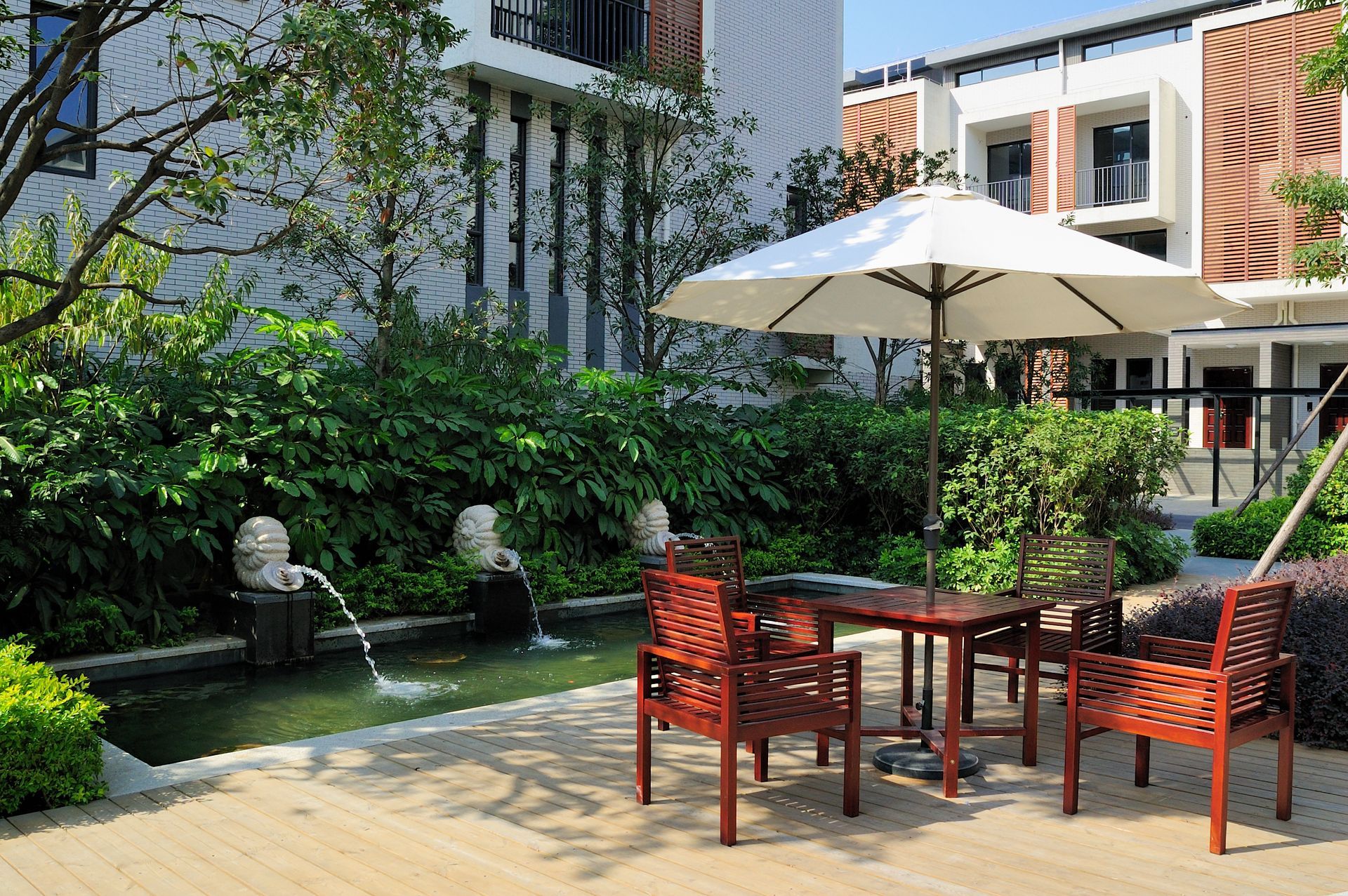 Outdoor patio with table, chairs, umbrella, and a water feature with greenery, next to a building.