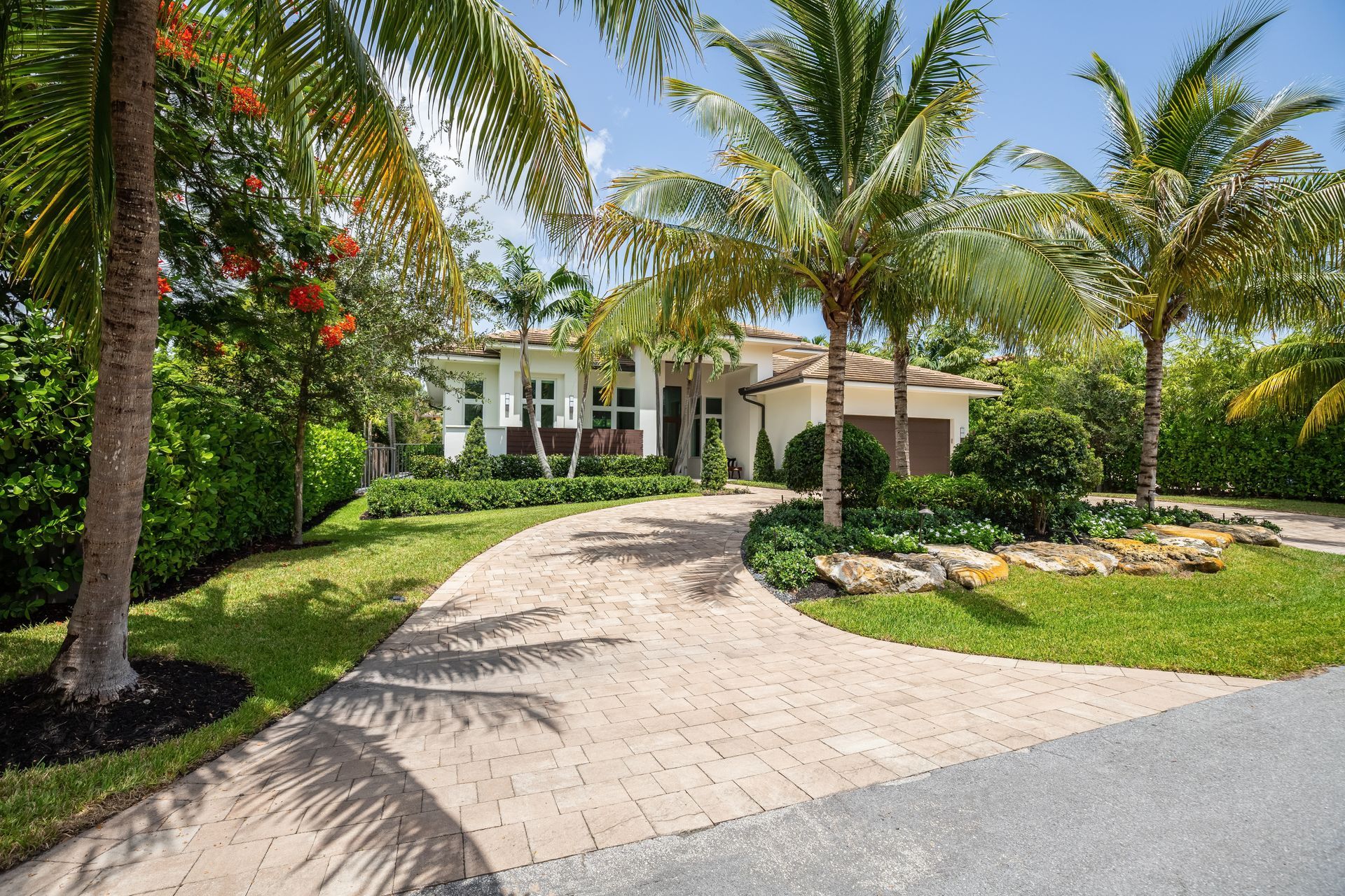 Paved driveway leading to a cream-colored house, framed by palm trees and lush green landscaping.