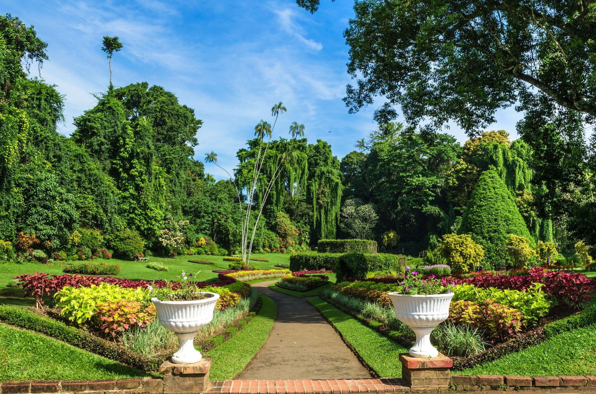 Formal garden with manicured hedges, flower beds, and a central path, under a bright blue sky.