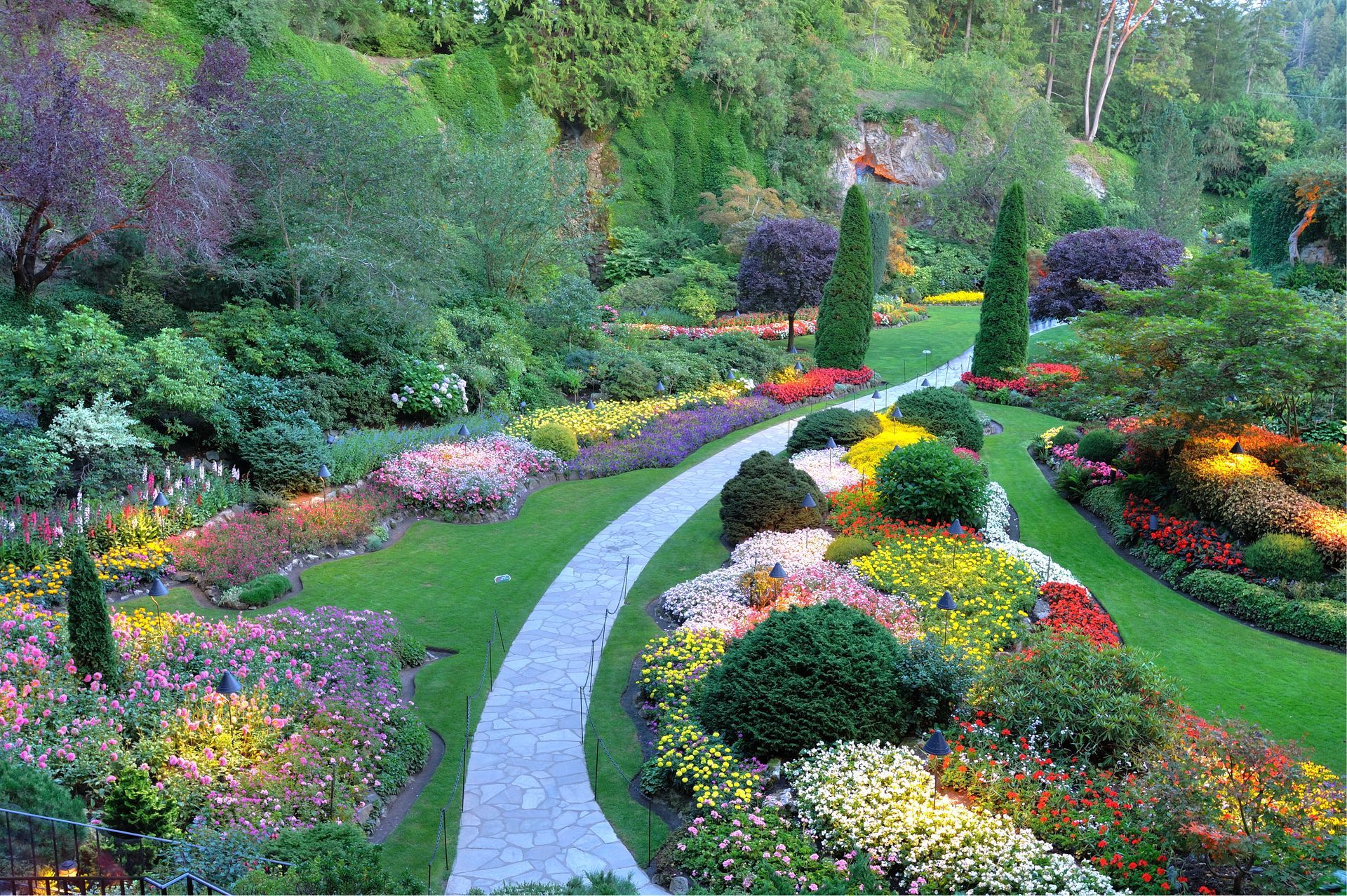 Lush garden with winding stone path, flanked by vibrant flower beds and green lawns under a bright sky.