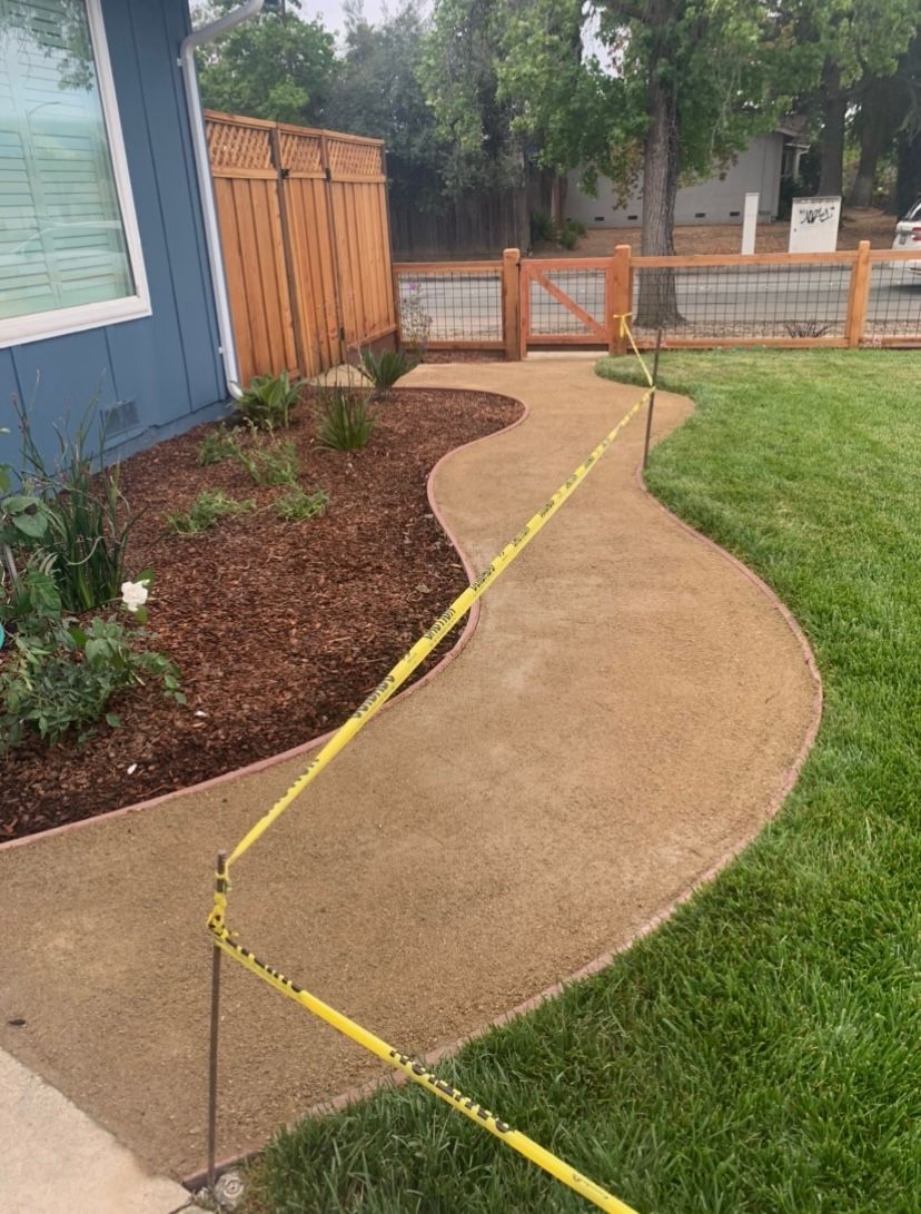 A newly paved tan walkway, bordered by grass and landscaping, leads to a gate in a wooden fence.