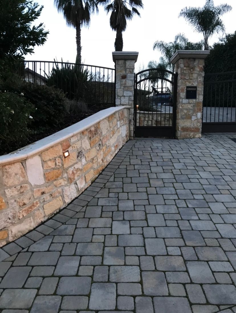 Stone driveway curving towards gated entrance; stone pillars, palm trees in background.