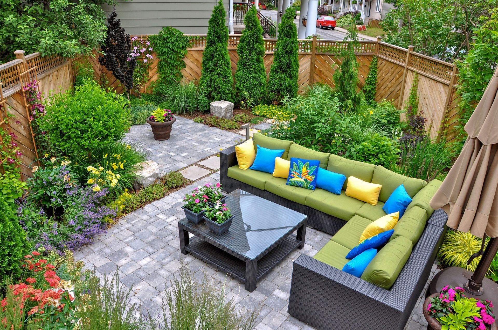 Cozy backyard patio with green sectional sofa, small table, and colorful pillows surrounded by lush greenery and a paved stone floor.
