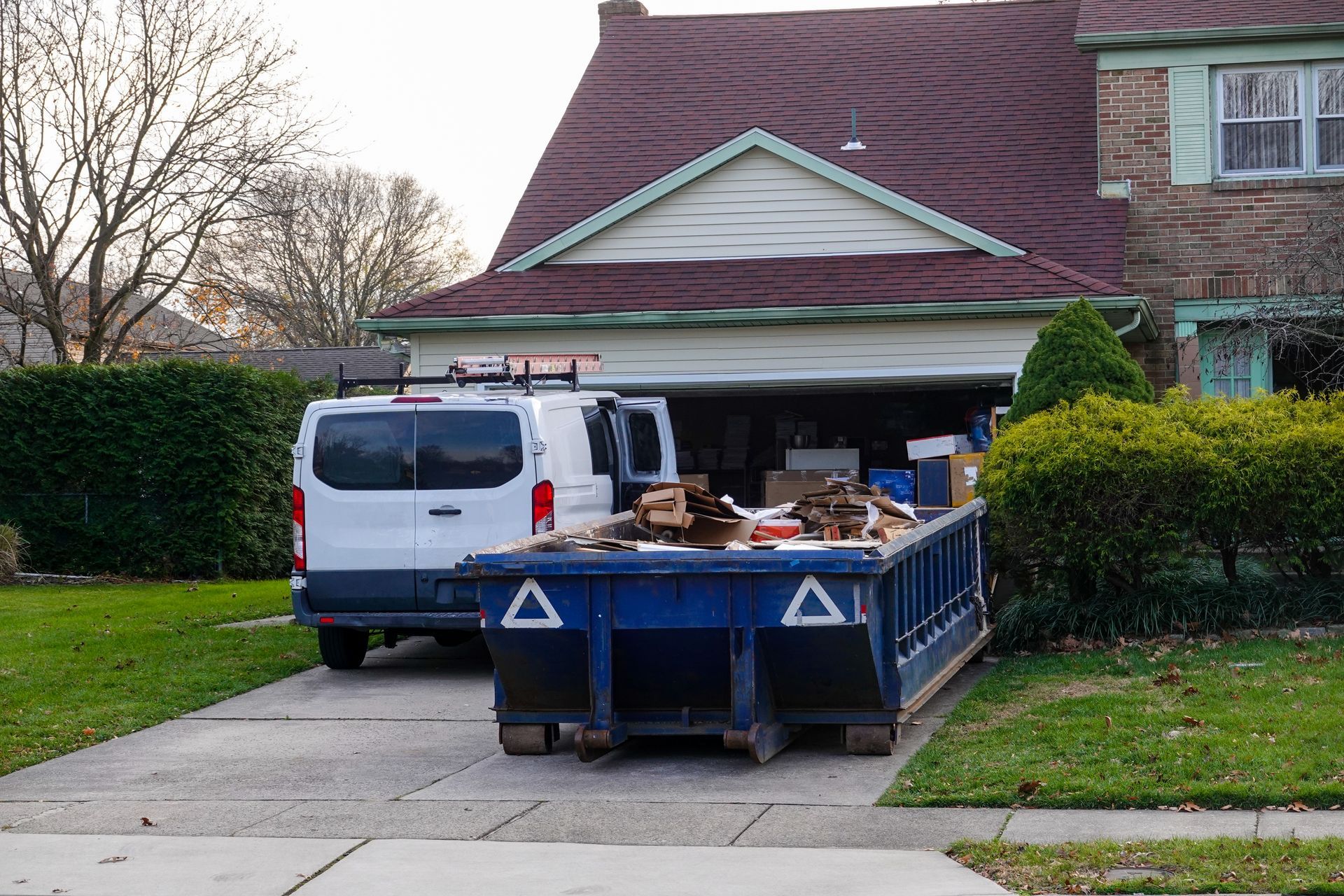 Blue dumpster in driveway with white van, garage open, residential yard.