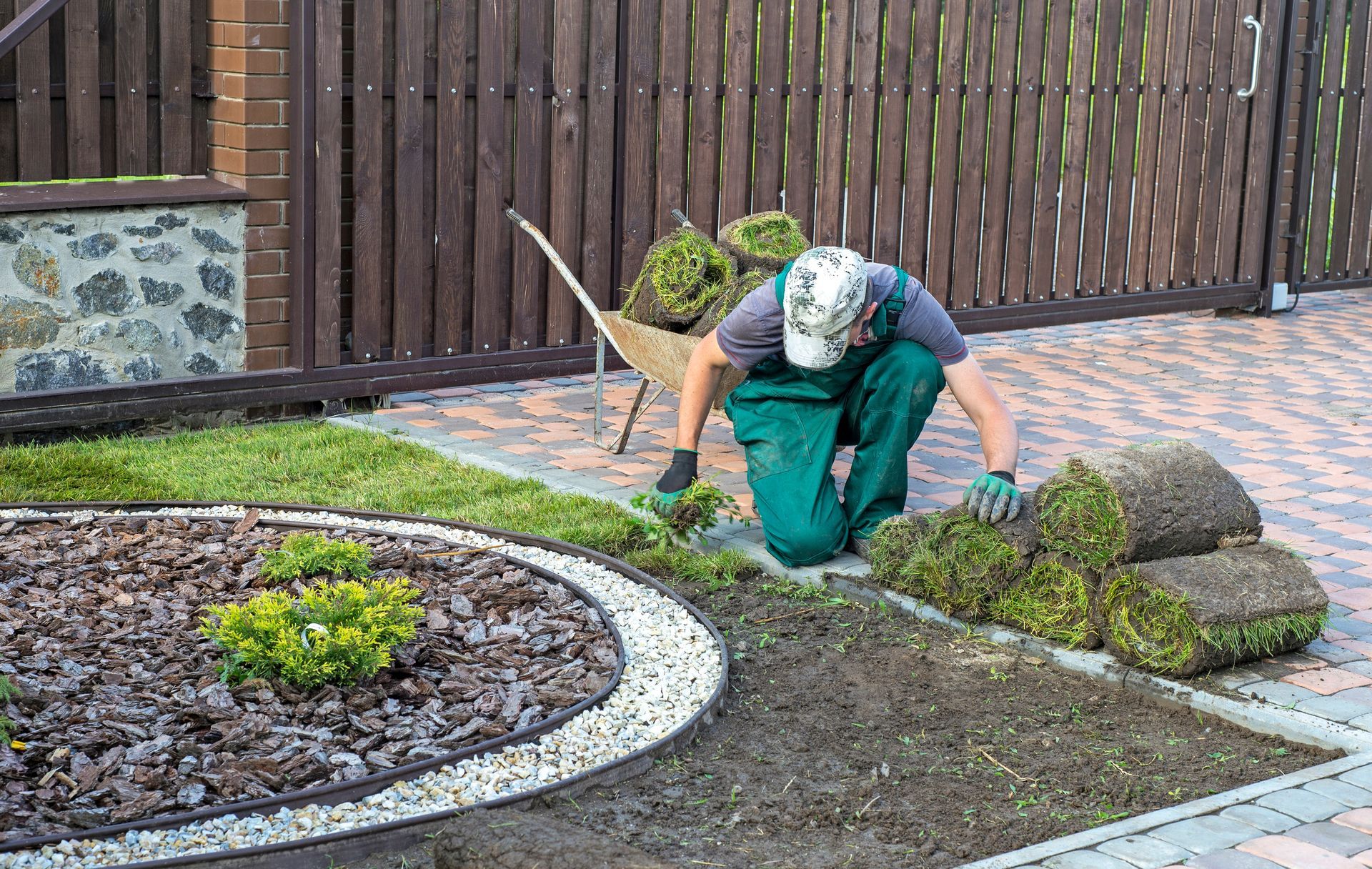 Man in green overalls laying sod in a garden bed next to a patterned driveway.