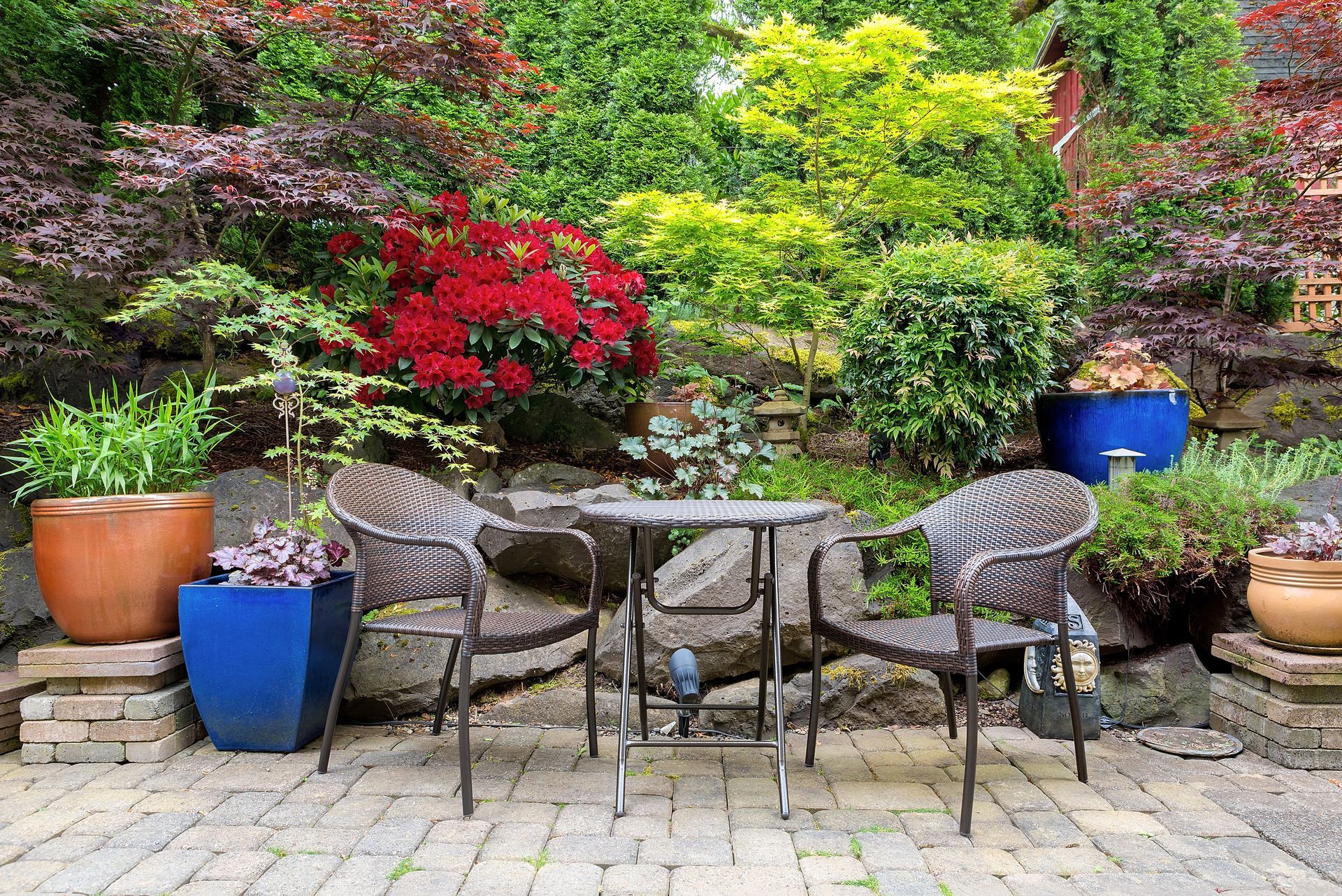 Patio seating area with table, chairs, colorful potted plants, and lush garden backdrop.