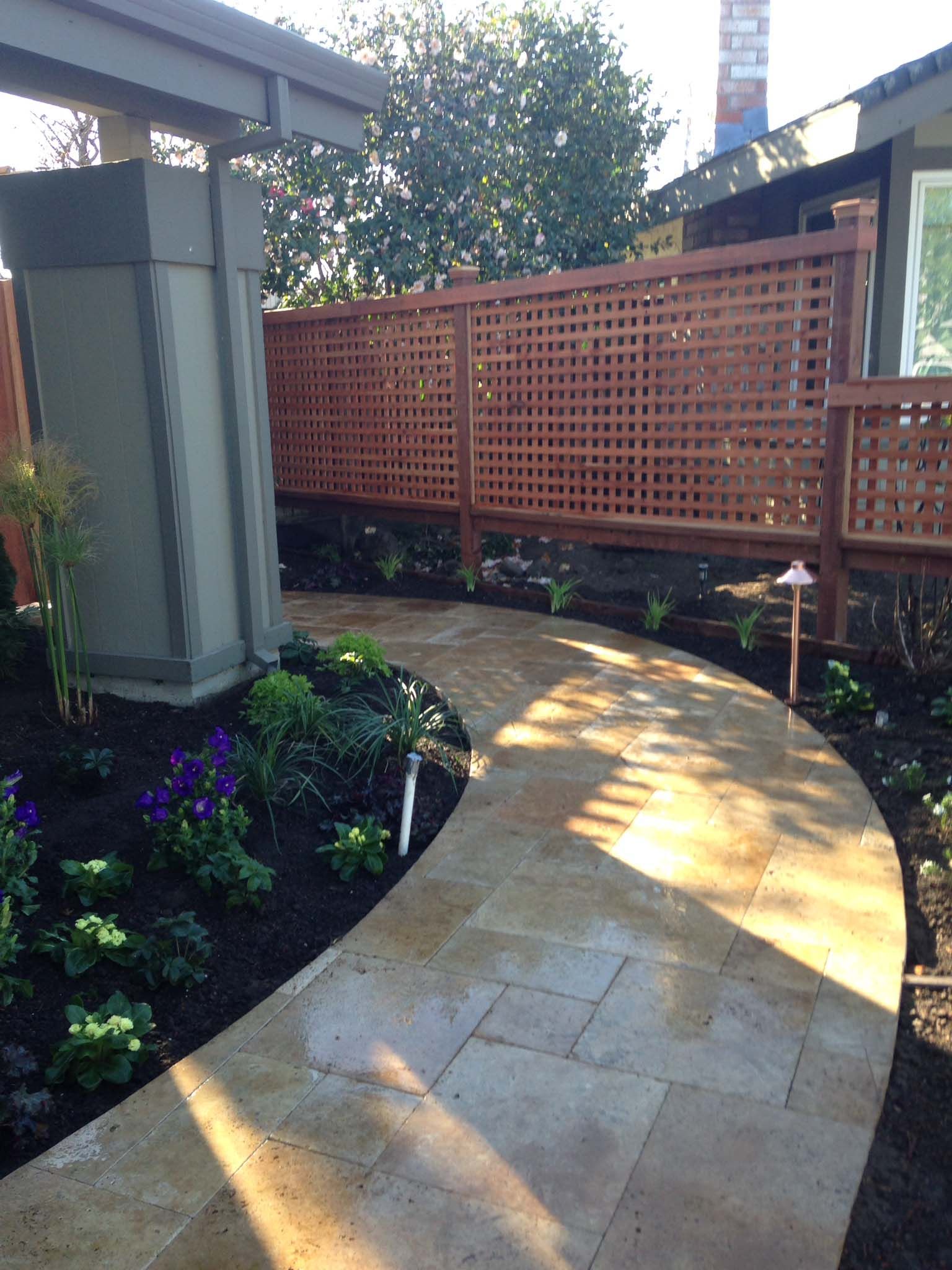 Stone pathway curves through a garden with a wood lattice fence, plants, and a gray post.