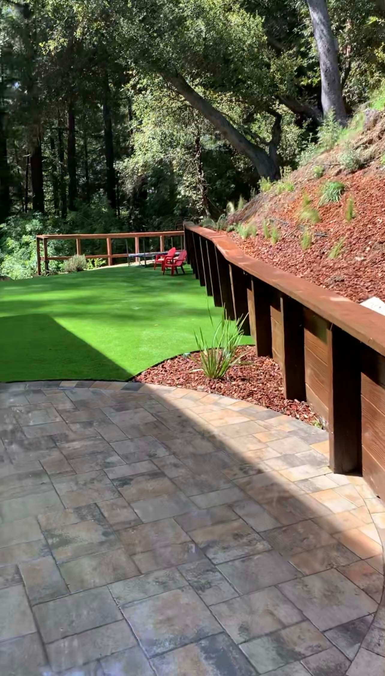 Patio with tiled floor, artificial grass lawn, wooden retaining wall, and forest backdrop.