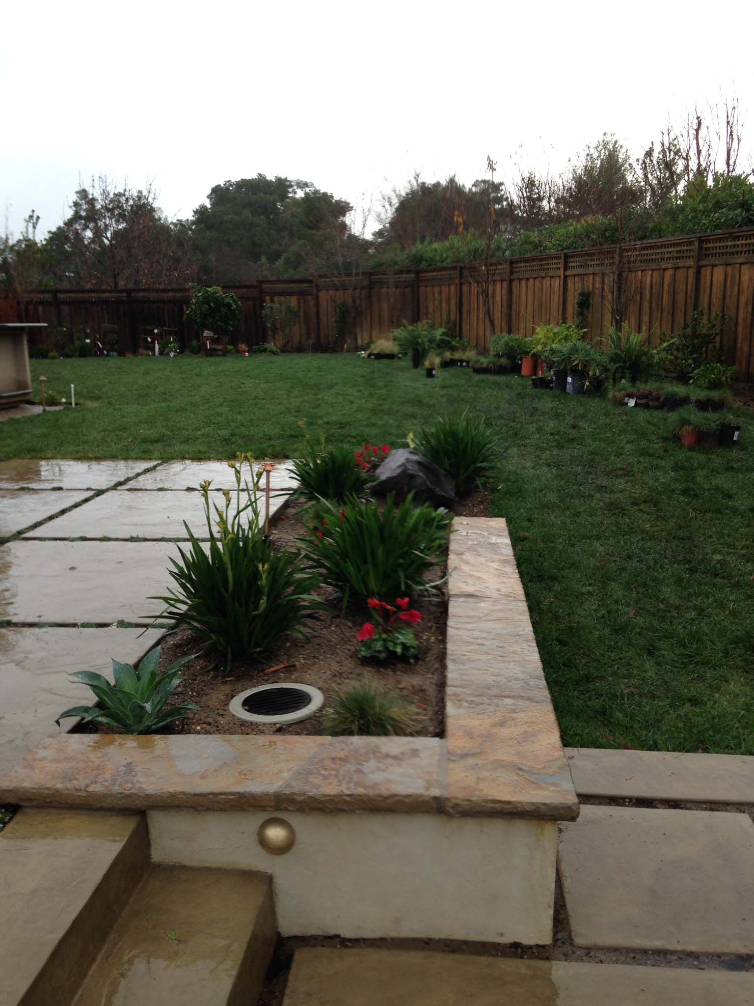A backyard with a stone patio, raised garden bed with flowers, and green lawn. A wooden fence is in the background.