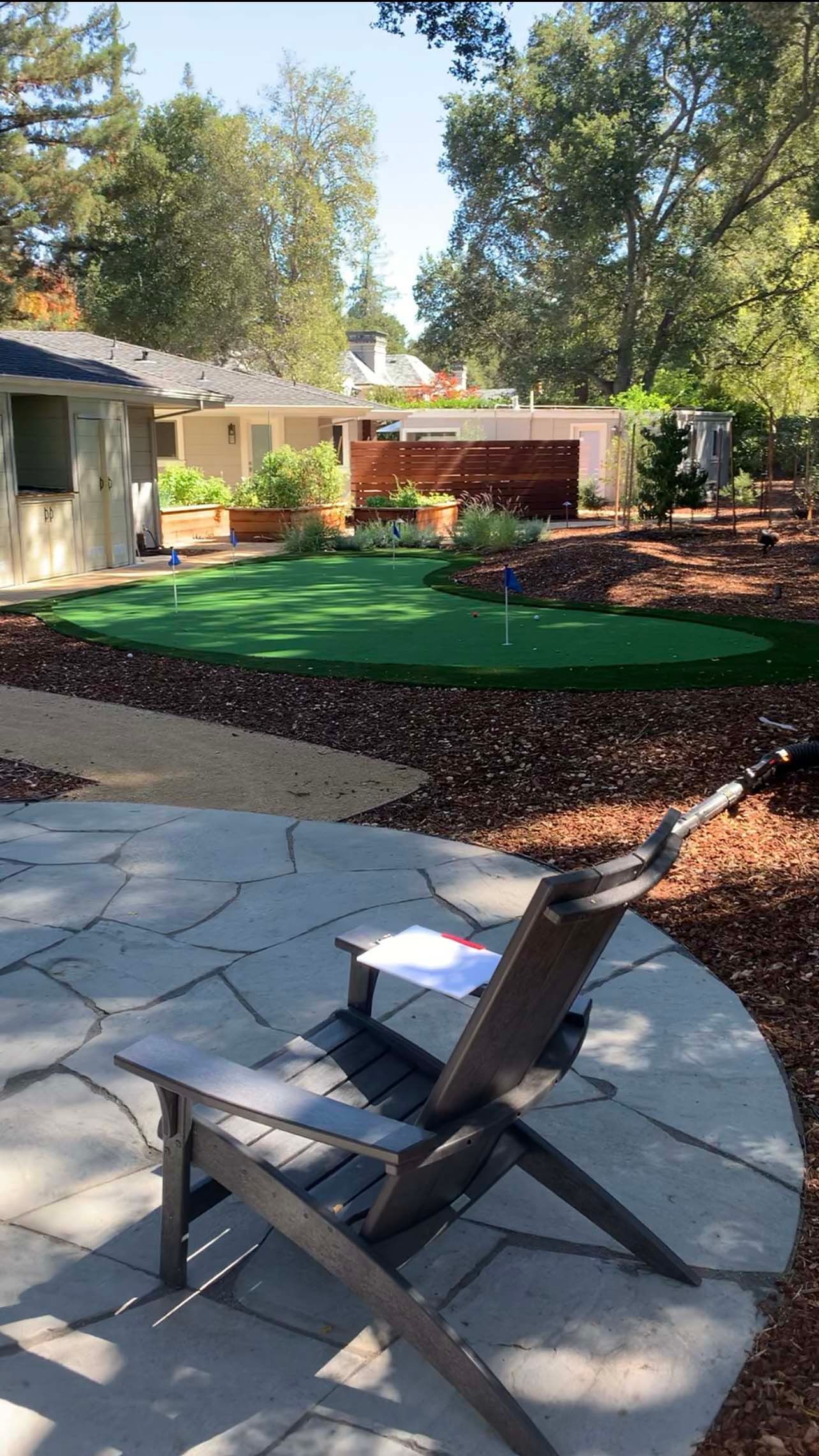 Wooden chair on stone patio overlooking a backyard putting green and house. Sunny day.