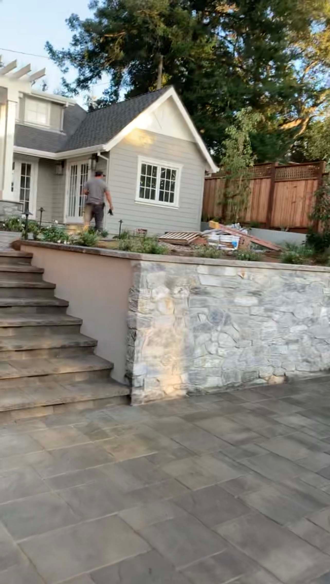 Stone retaining wall with steps leading to a gray house, person walking, paved patio.