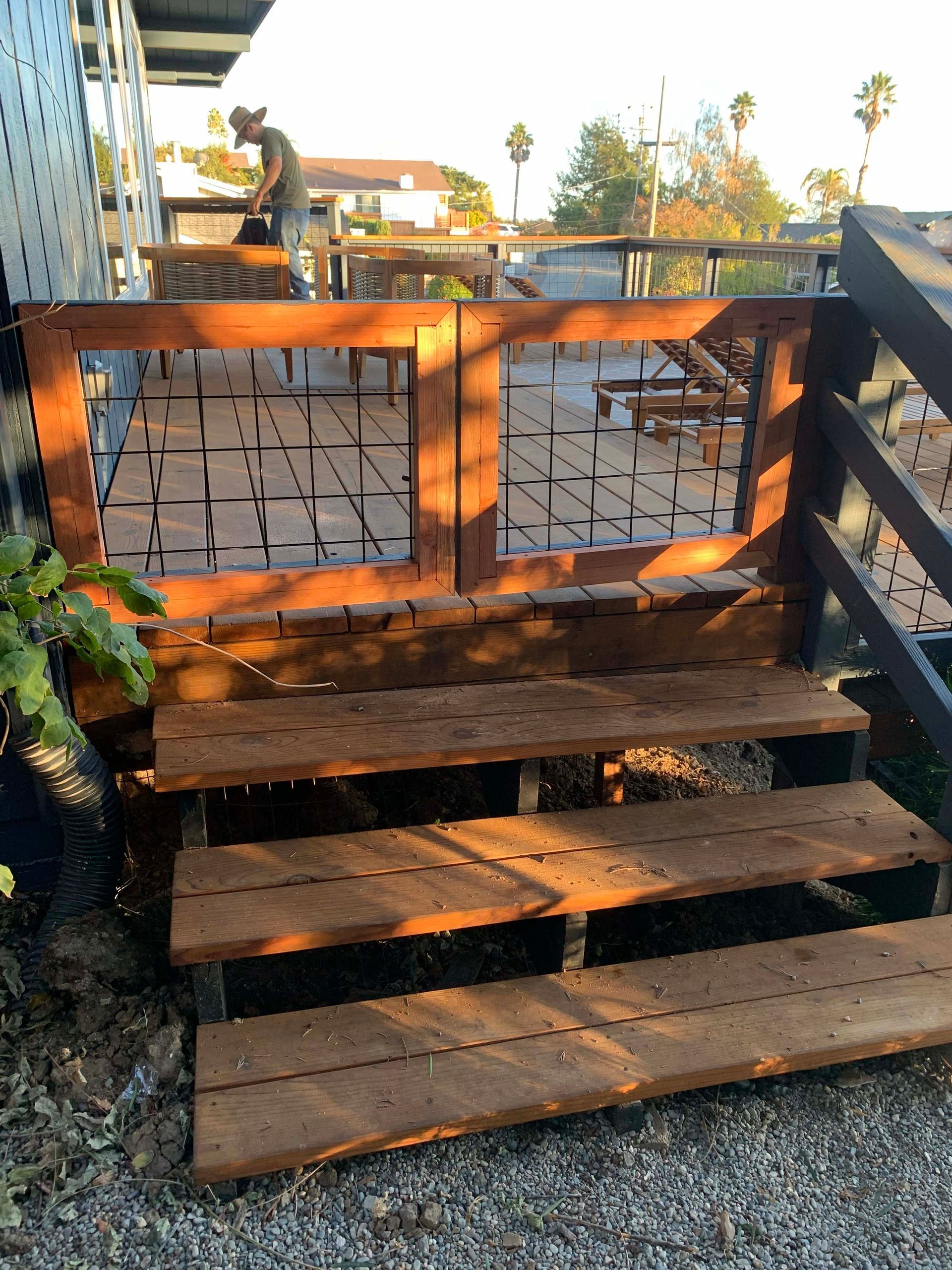 Wooden steps leading up to a patio with a decorative gate; person working in the background.