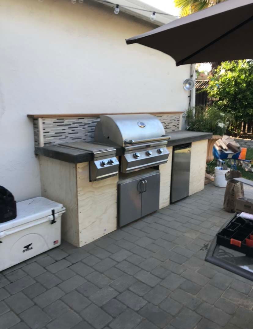Outdoor kitchen with built-in grill, refrigerator, and side burner on a paved patio next to a white wall.