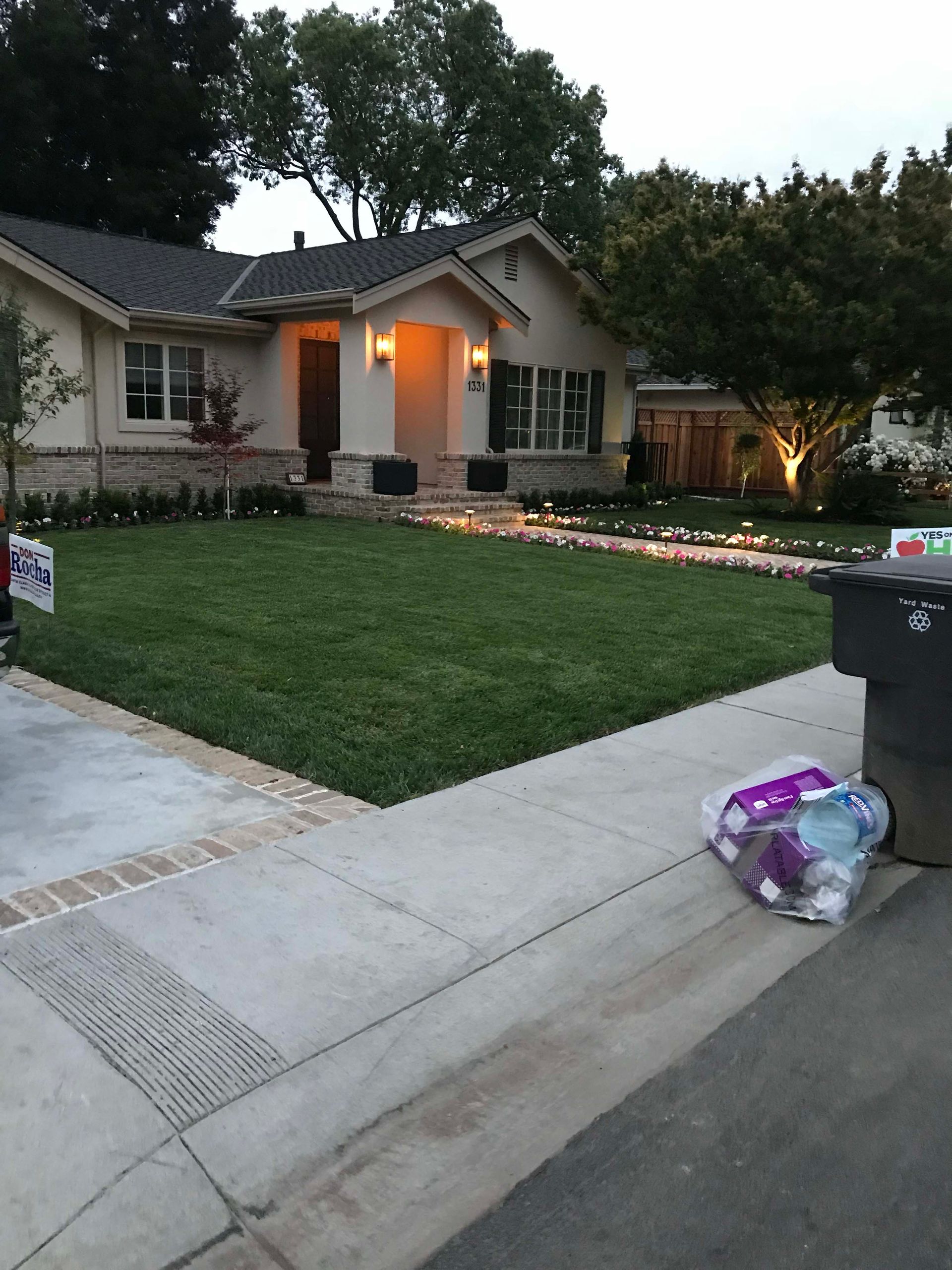 House with green lawn, sidewalk, and trash cans. Evening scene, warm lighting.