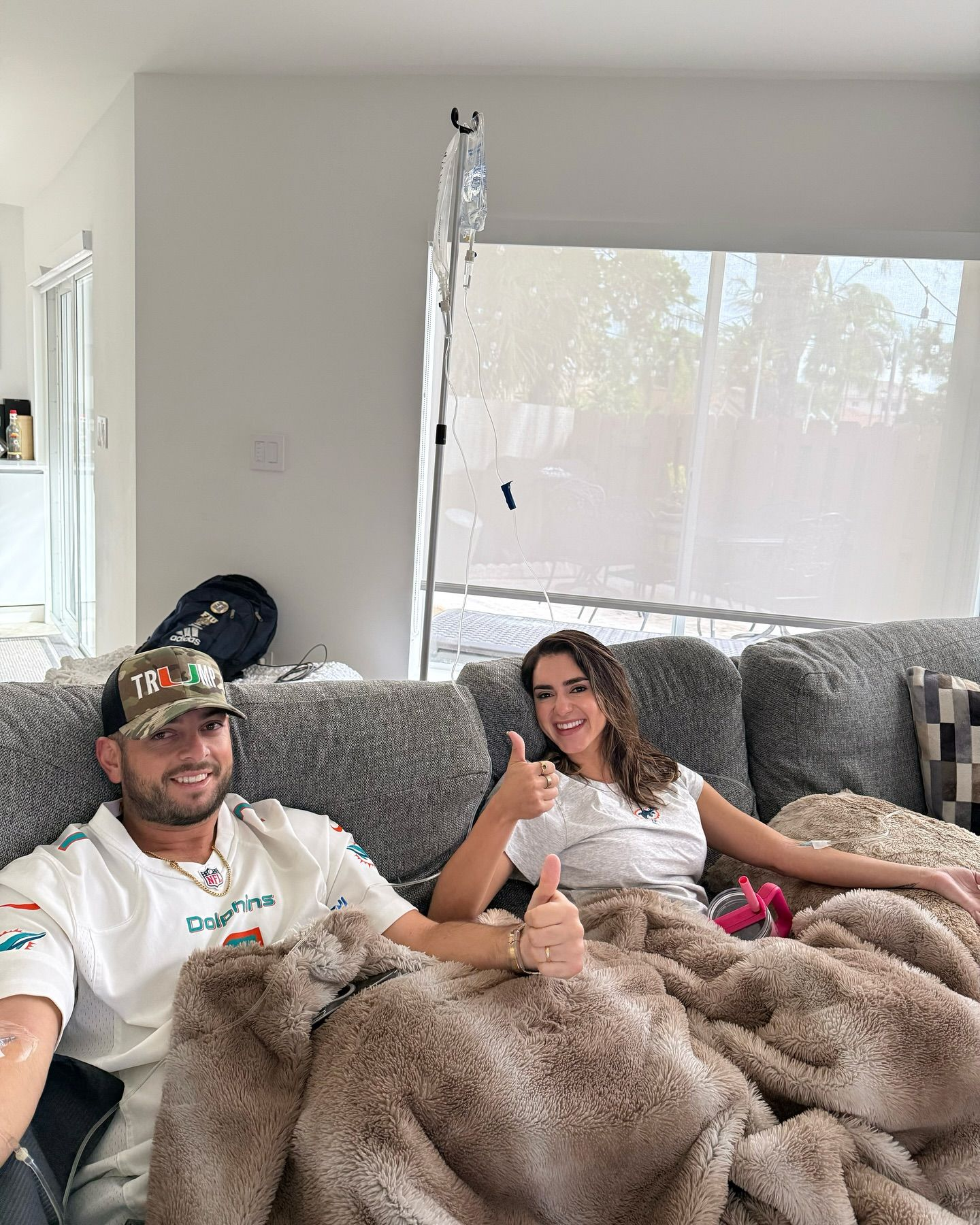 Couple on a couch, covered by a blanket, giving thumbs-up. Bright room with window.