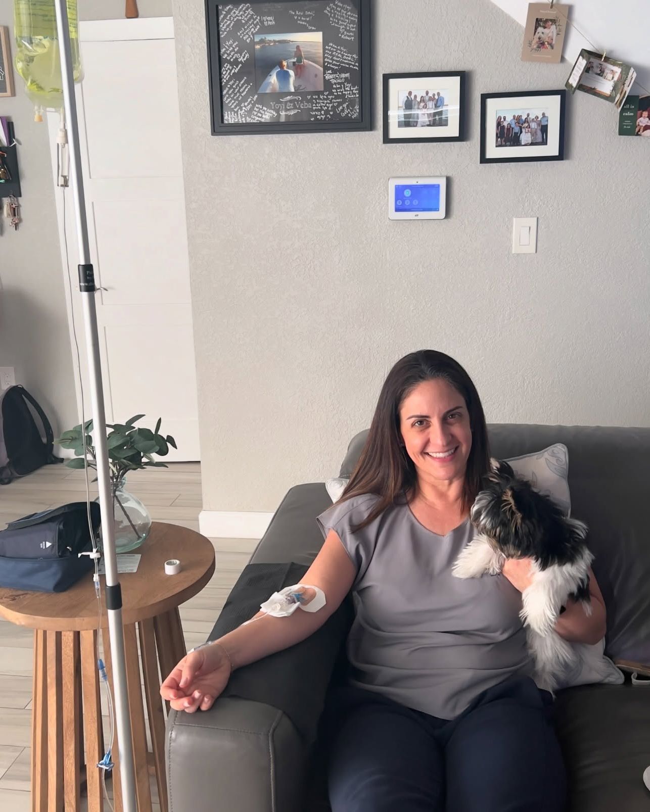 Woman on couch receiving IV treatment, holding a small dog. Room setting with photos on the wall.