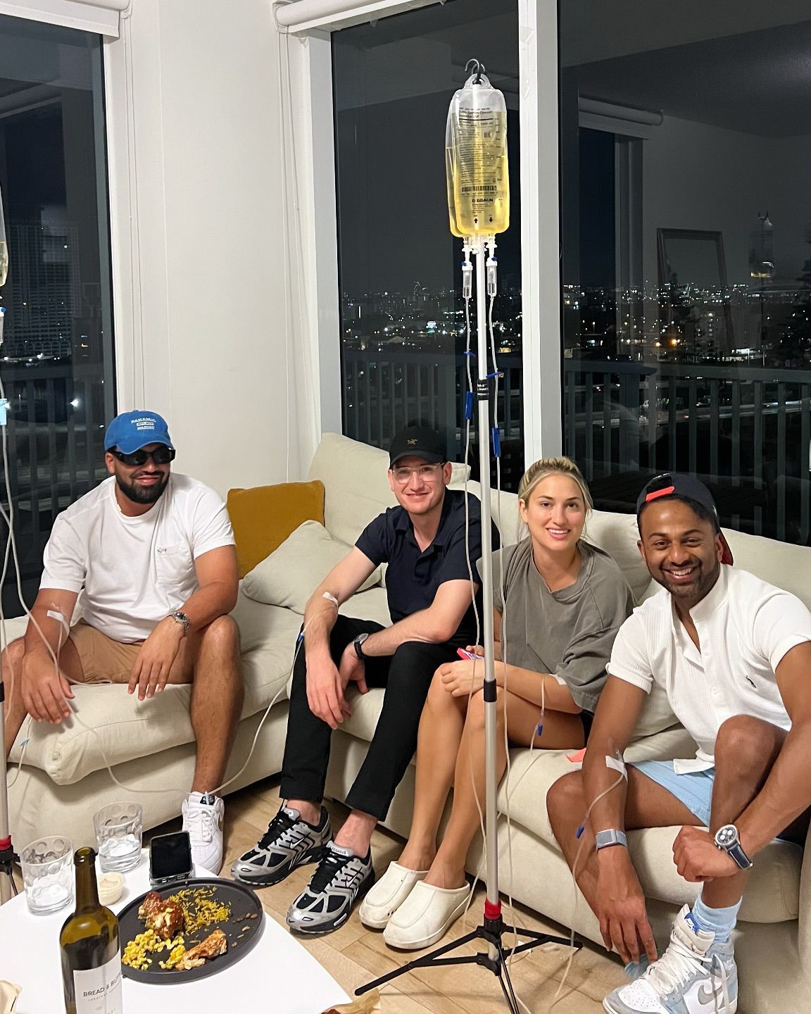 Four people receiving IV drips while sitting on a couch. The setting appears to be a modern apartment with city views.