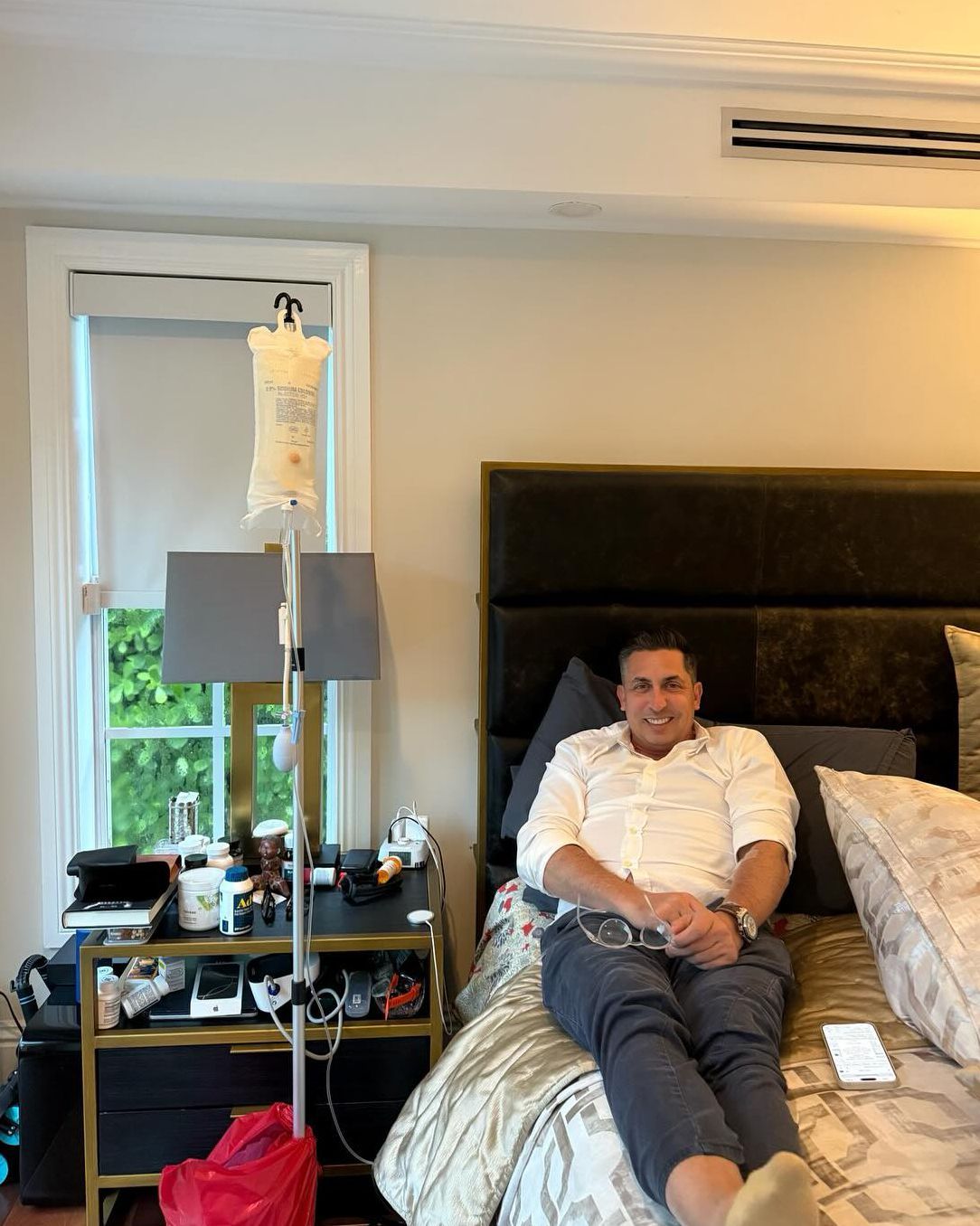 Man receiving IV fluids, sitting on bed, smiling. Bedroom setting with window, medical equipment visible.