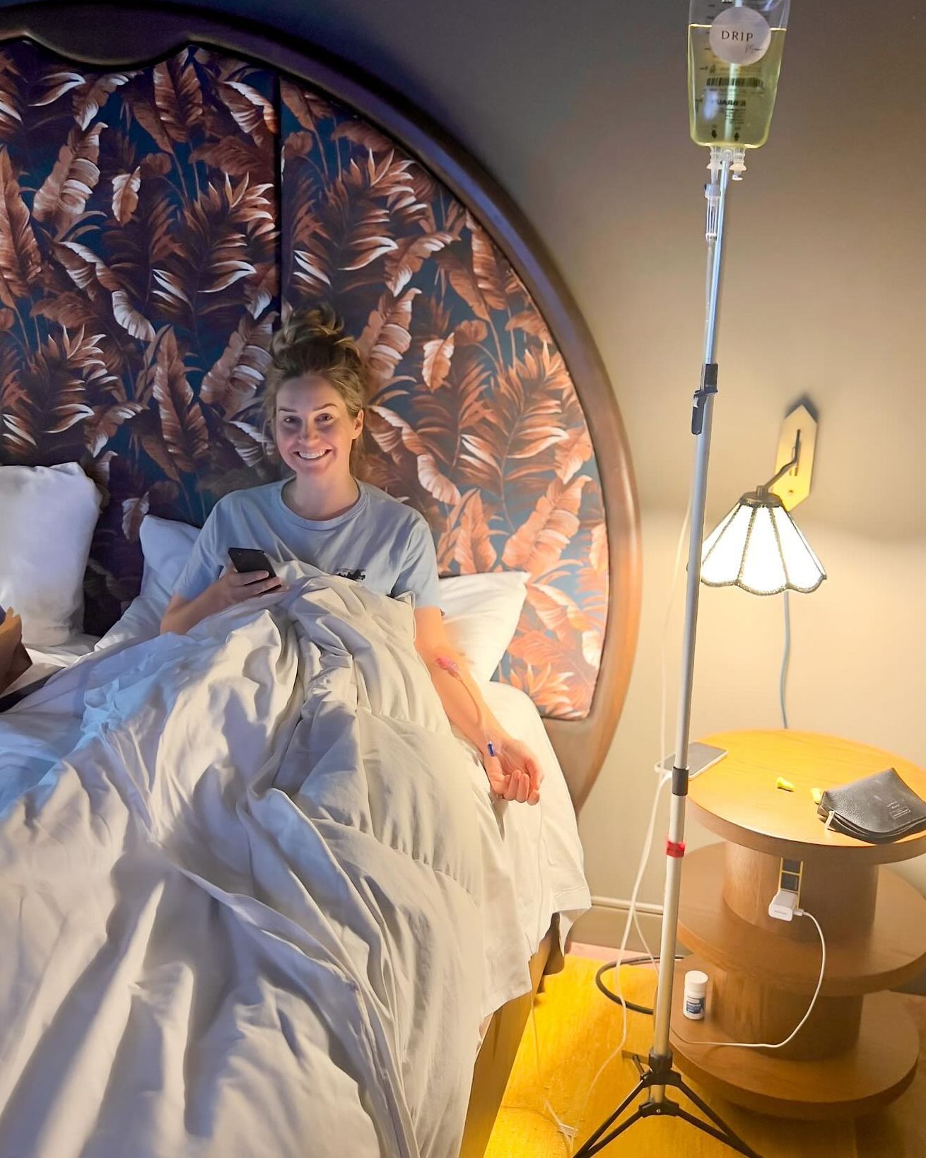 Woman in bed receiving IV fluids, smiling at phone. Bedside table, floral headboard, and infusion stand.
