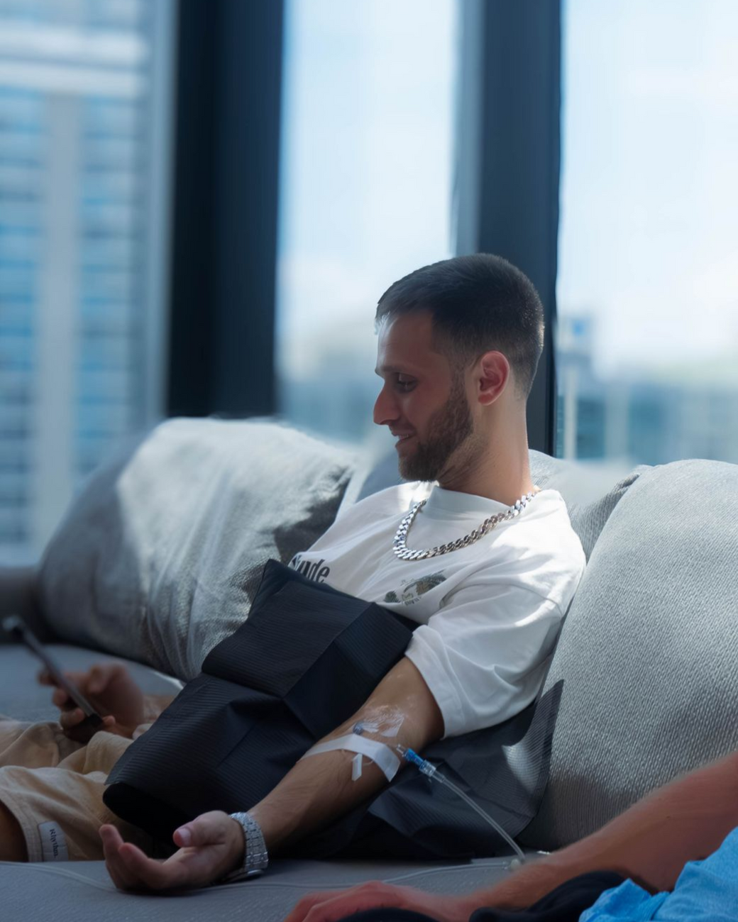 Man on couch with IV drip in arm; smiles; window background.
