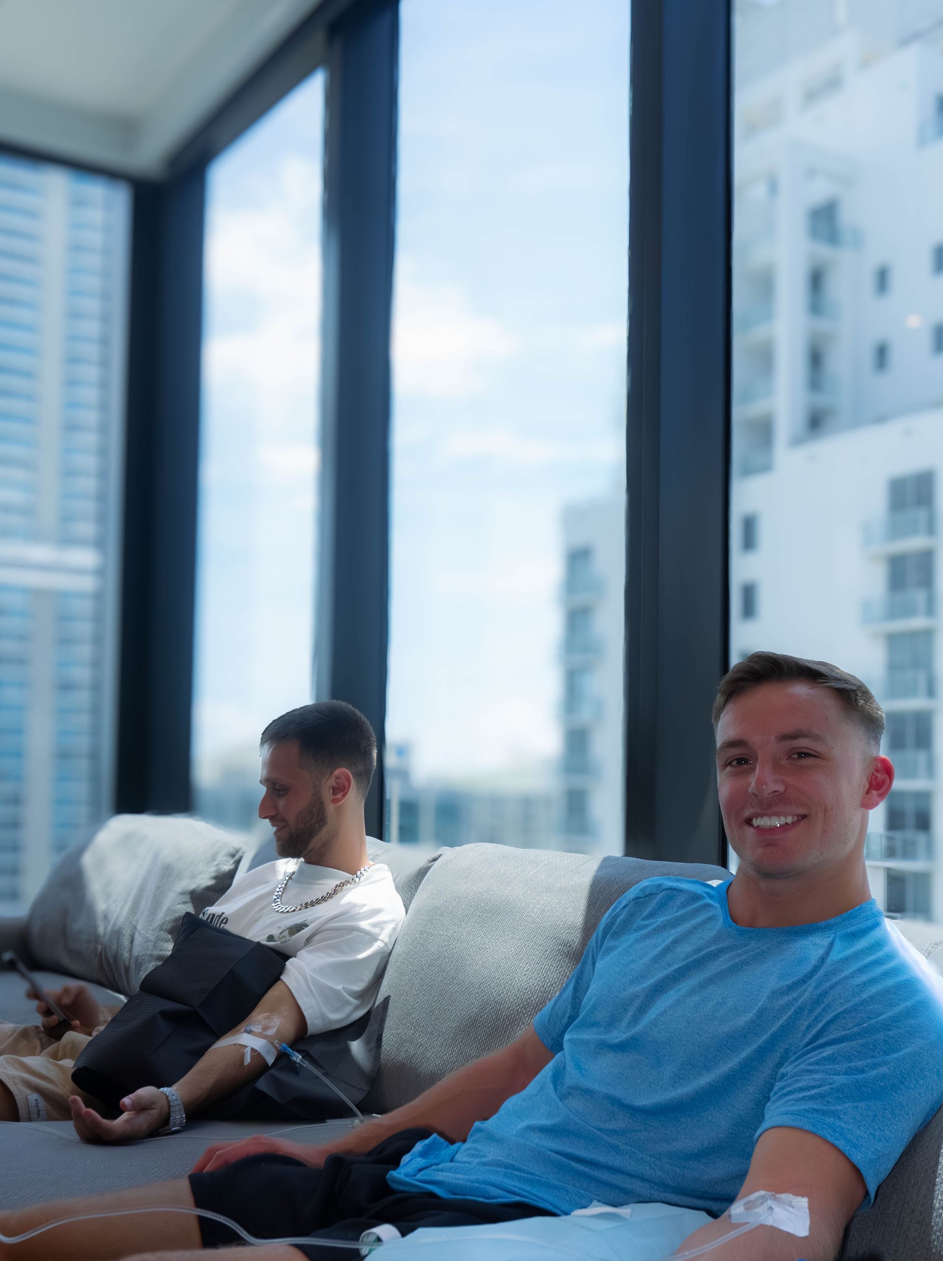 Two men receiving IV drips on a couch by a window; one smiles, the other looks away. Sunny day outside.