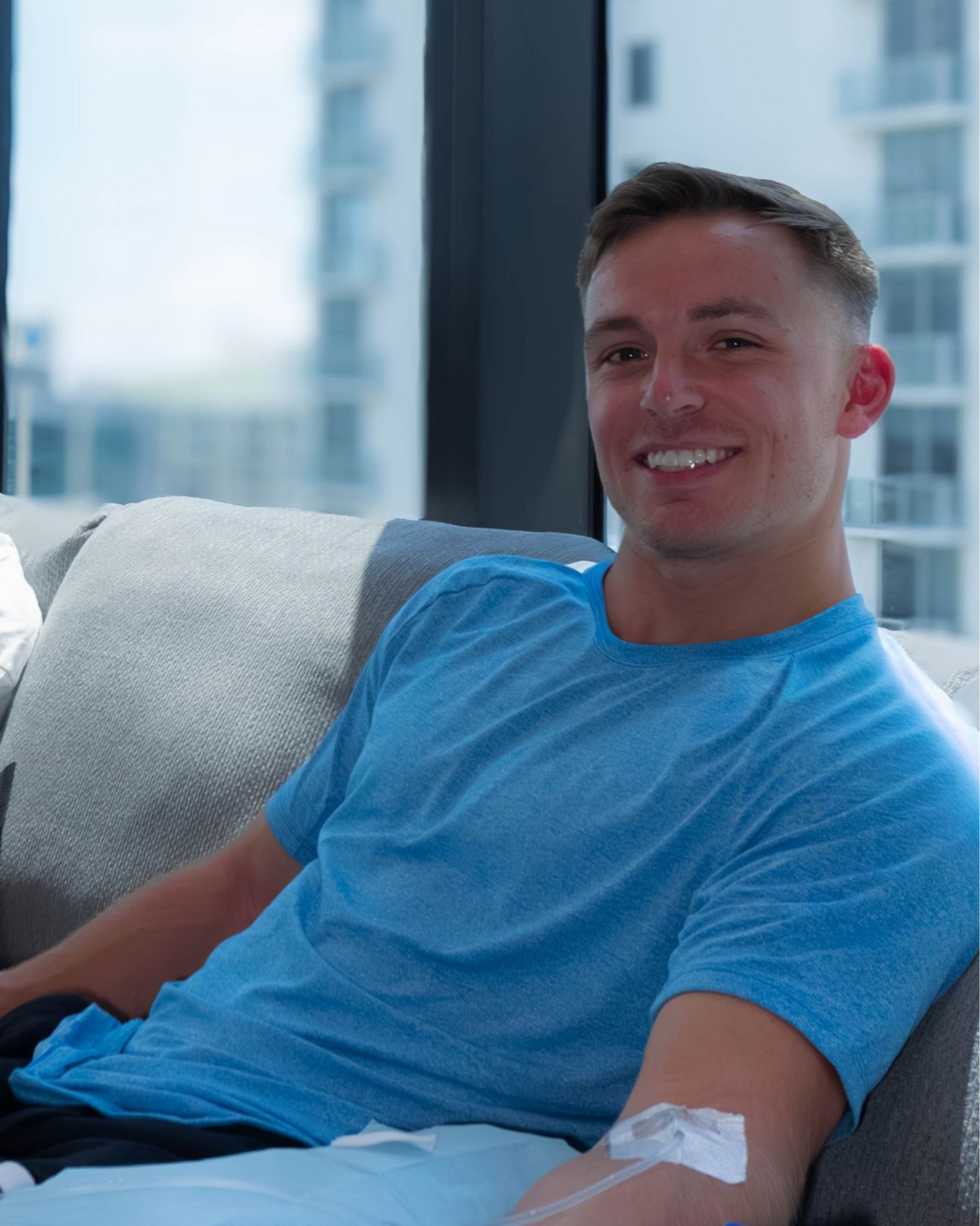 Man wearing a blue shirt smiles while receiving an IV treatment. Seated indoors with a cityscape view.